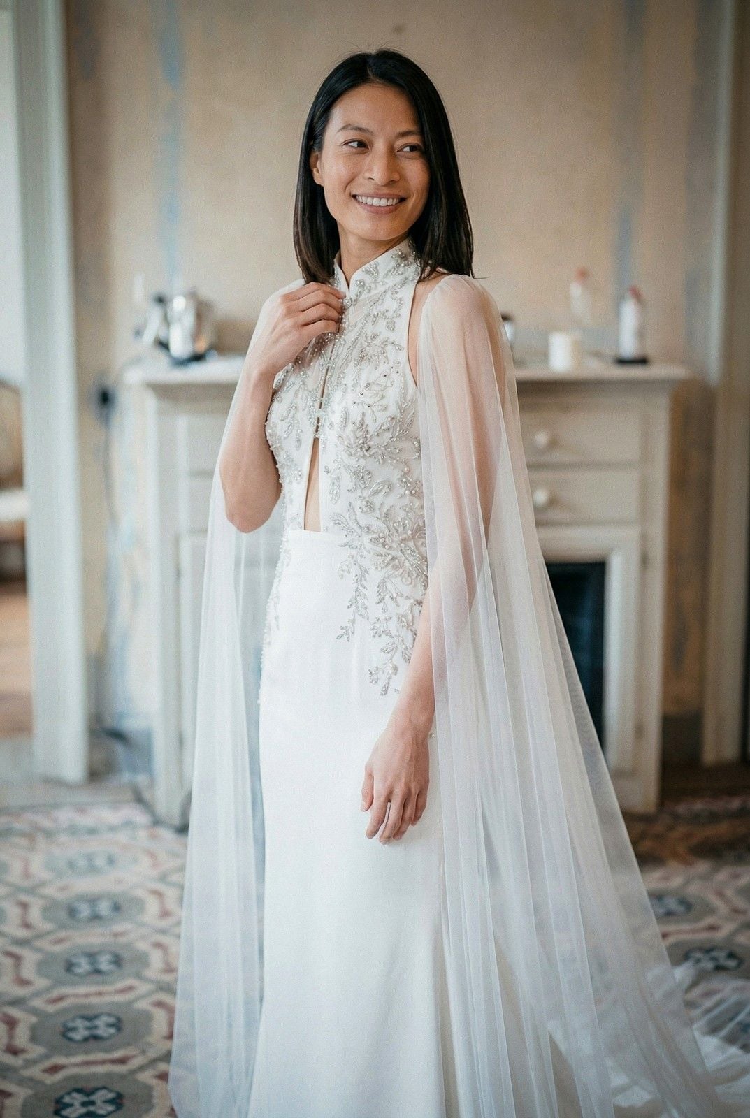 A woman in an elegant wedding dress with a sheer cape smiles warmly, showcasing intricate details and a charming setting.