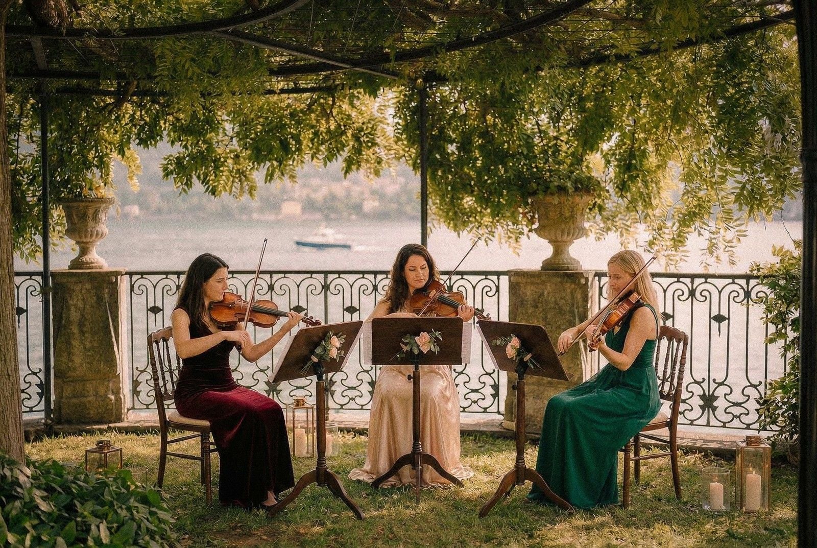 Three women in elegant dresses play violins under a canopy of greenery, with a serene lake in the background.