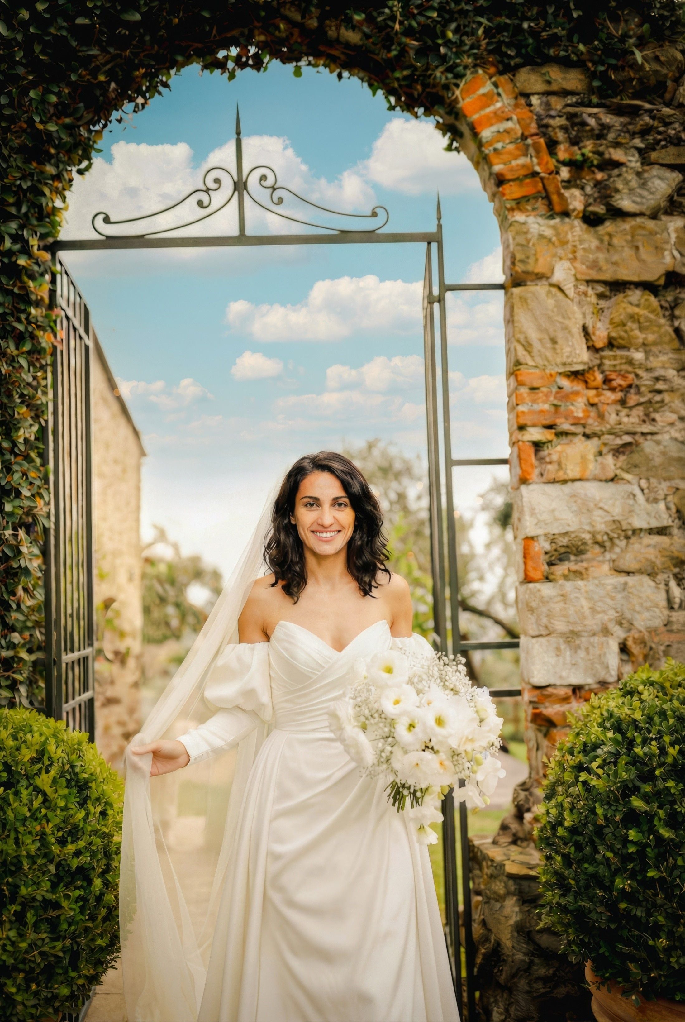Bride in a stunning white gown stands at the garden entrance, holding a bouquet of white flowers against a picturesque sky.