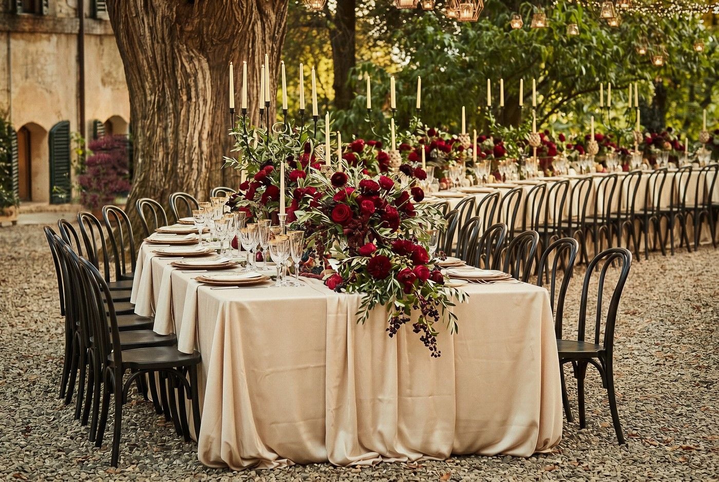 An elegant outdoor wedding setup features long tables adorned with lush red floral arrangements and candlelight.