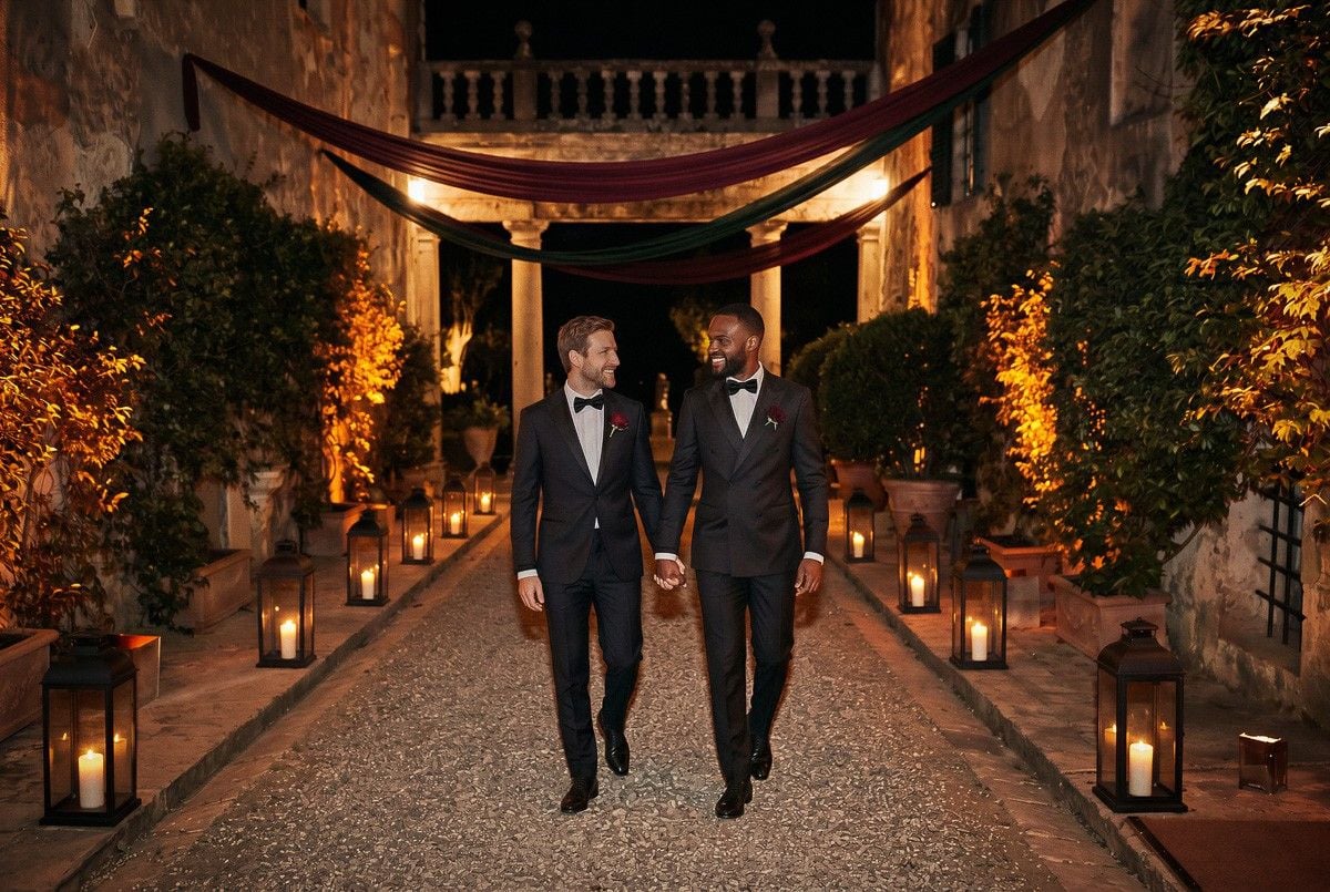 Two grooms dressed in tuxedos walk hand in hand down a romantic, candlelit pathway surrounded by greenery.