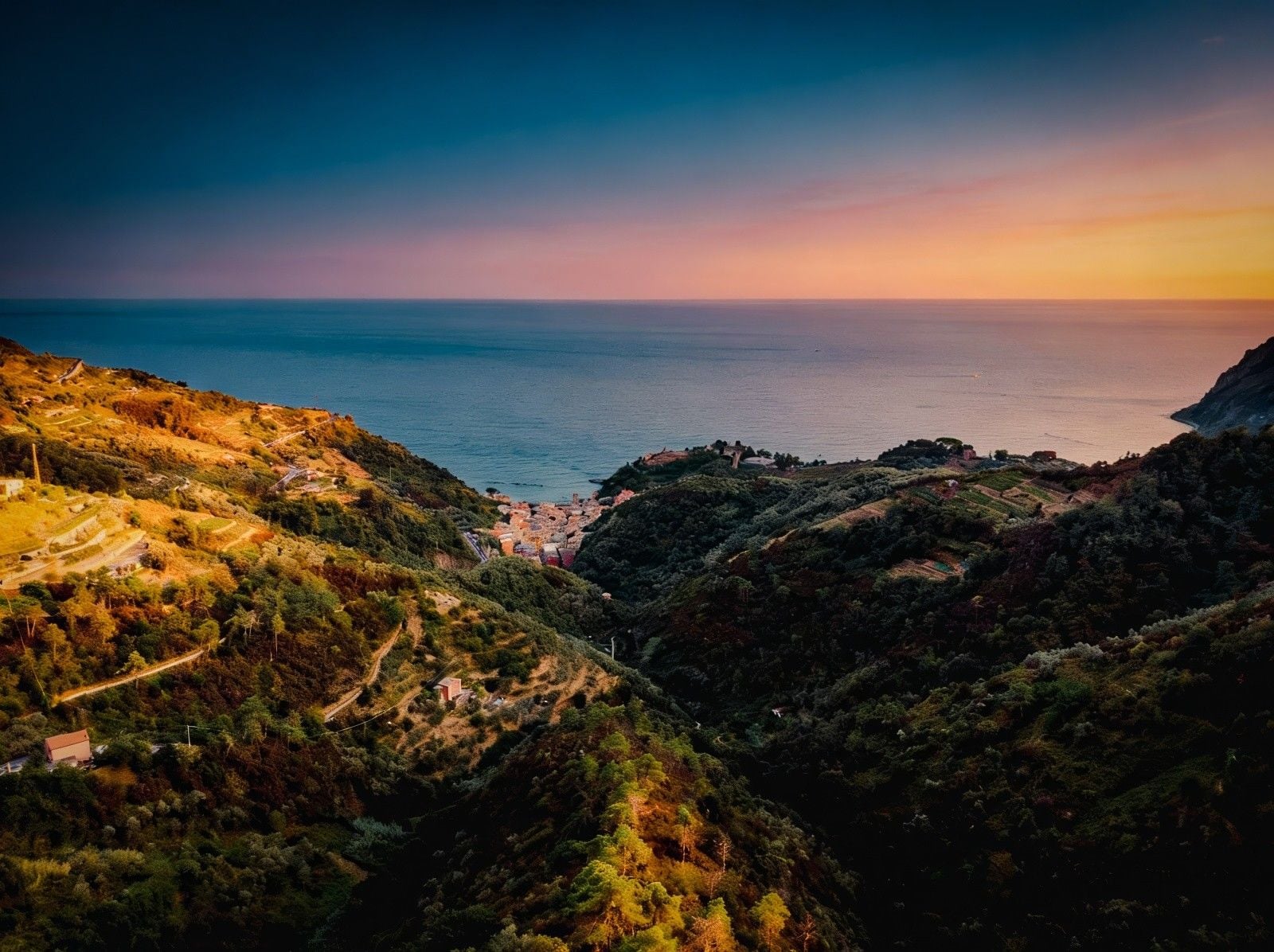 Sunset casts warm hues over hills leading to a coastal village, framed by lush greenery and the serene sea.
