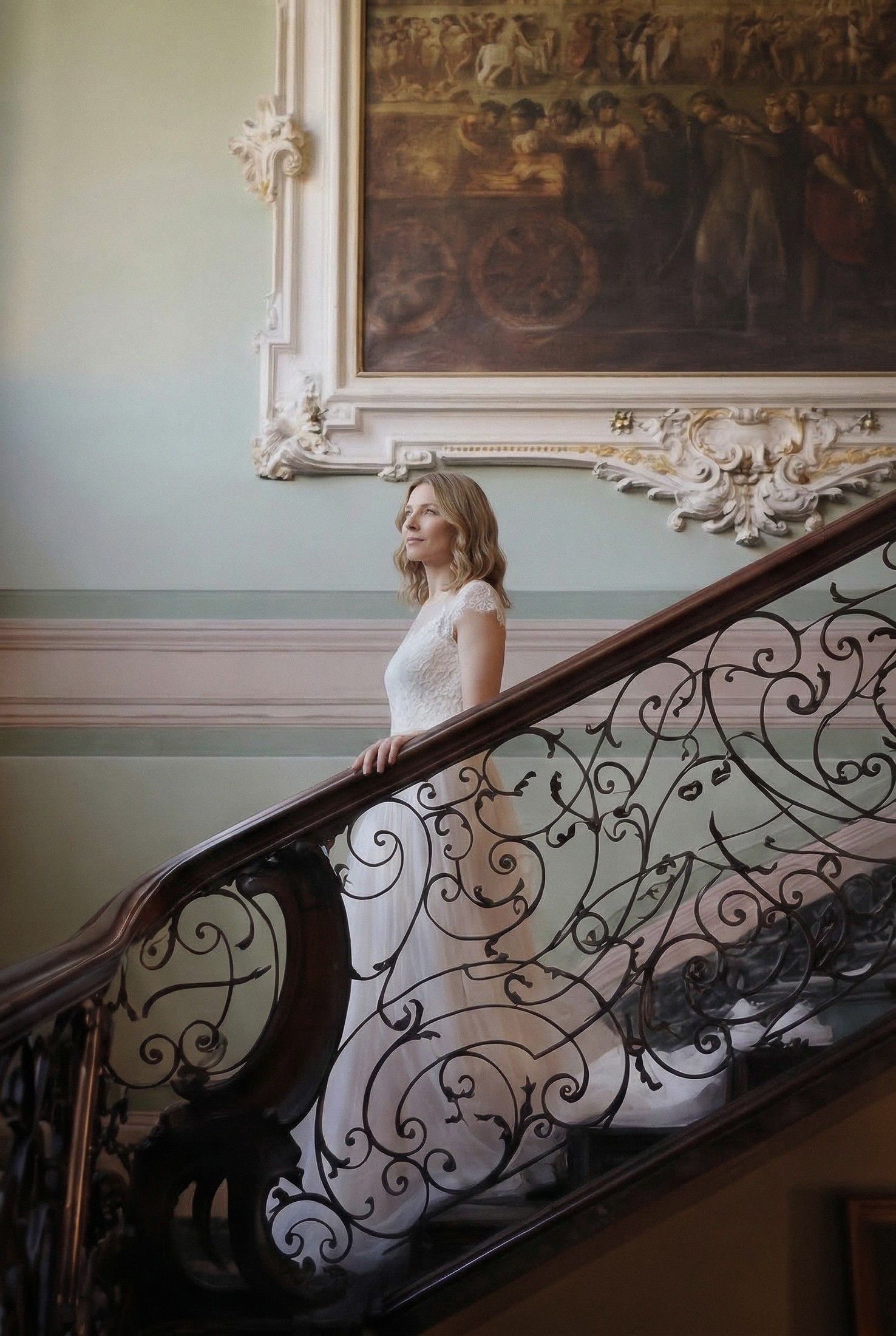 A bride in a flowing white gown stands gracefully on a staircase, illuminated by soft light and ornate decor.