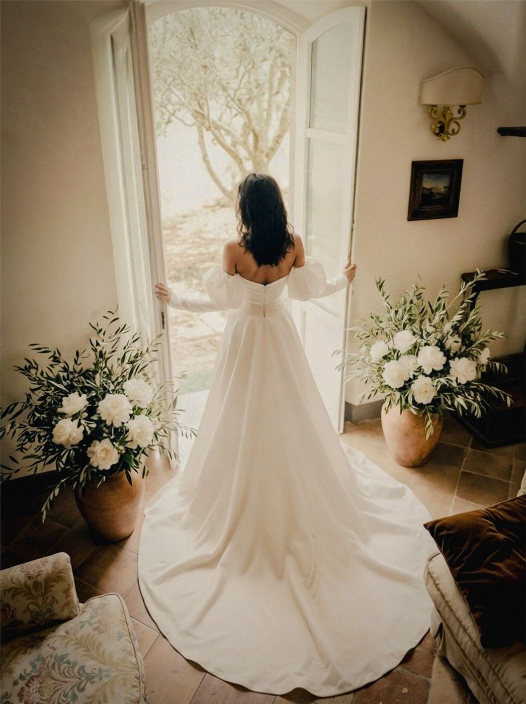 Bride in a flowing white gown stands at an open door, surrounded by lush floral arrangements and soft natural light.