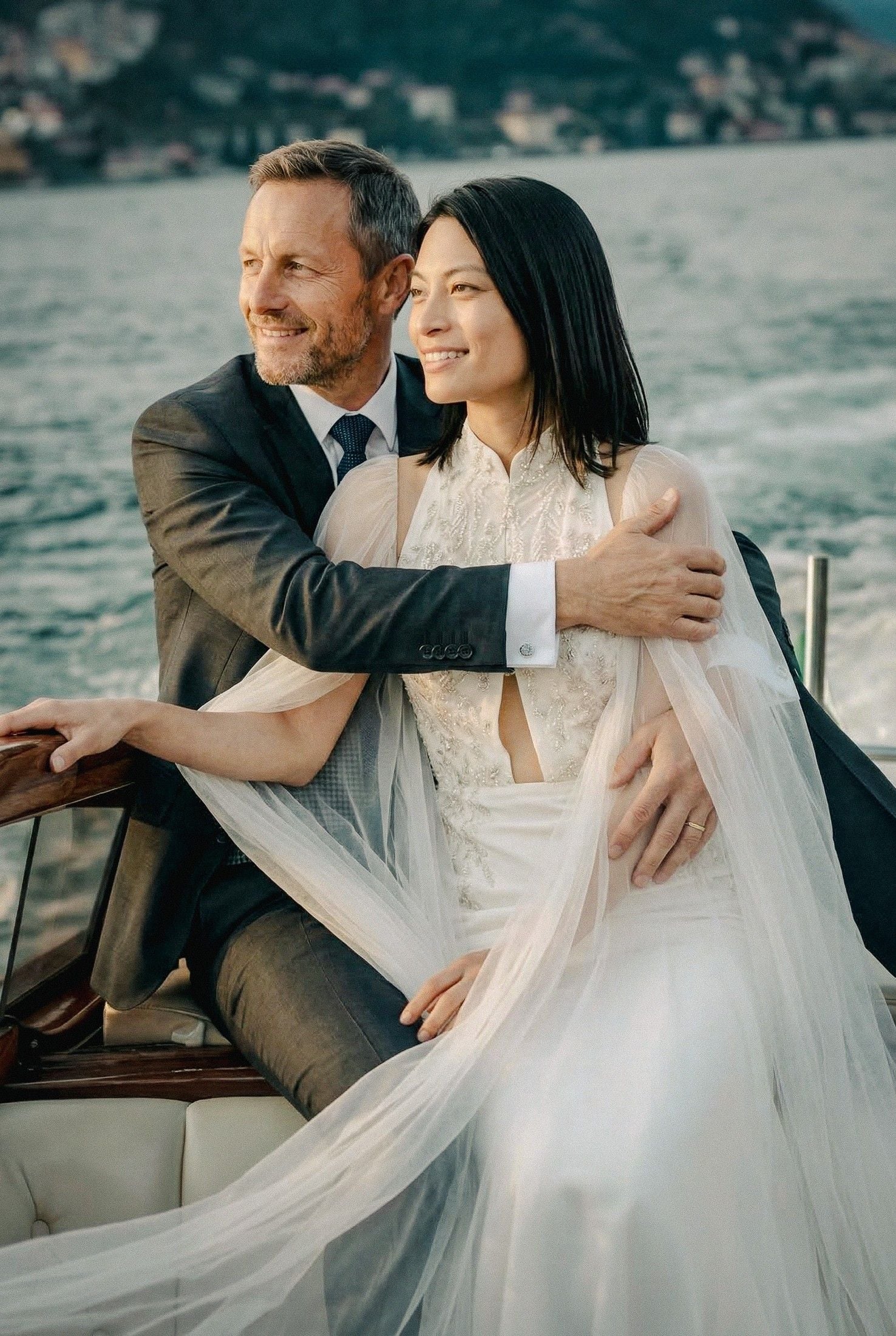 A joyful couple in elegant wedding attire embraces on a boat, with a shimmering lake and green hills in the background.