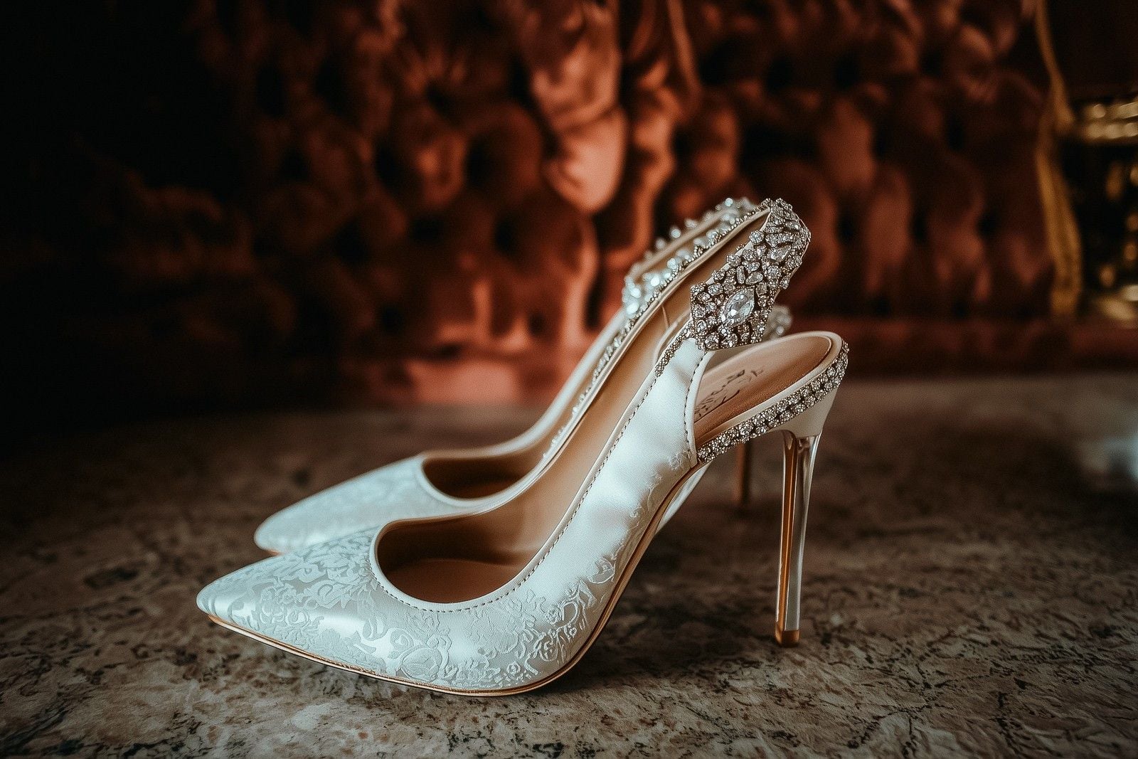 Elegant white bridal heels with intricate detailing, resting on a marble surface against a plush, dark backdrop.