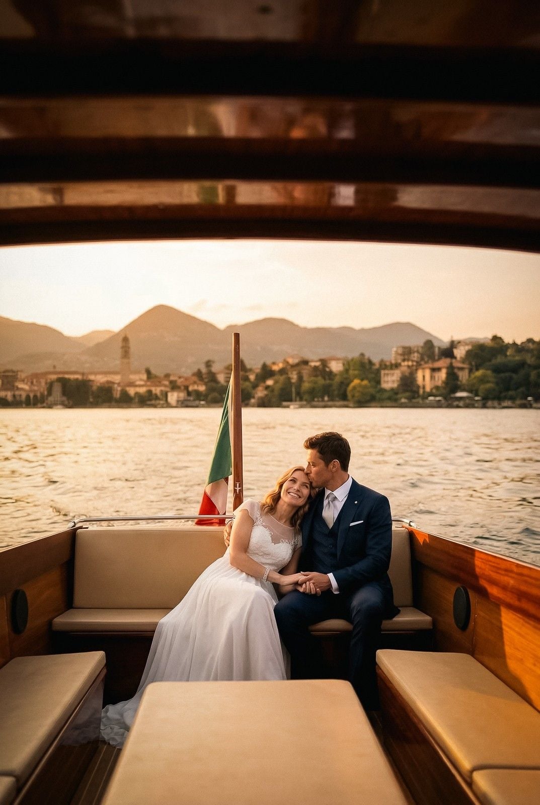 A couple in wedding attire sits on a boat, sharing a tender kiss with a picturesque lake and mountains in the background.