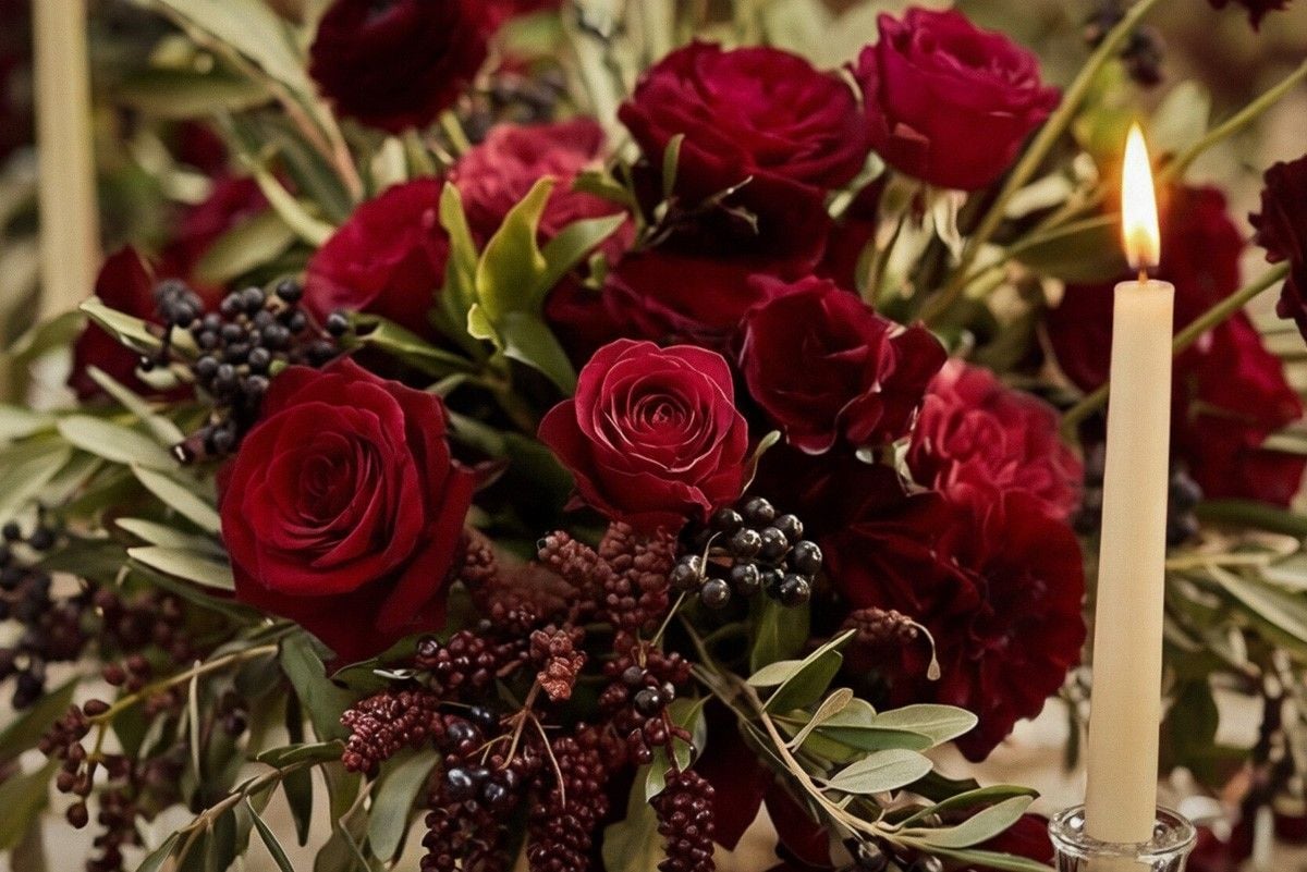 A romantic arrangement of deep red roses, greenery, and berries, with a lit candle creating a warm atmosphere.