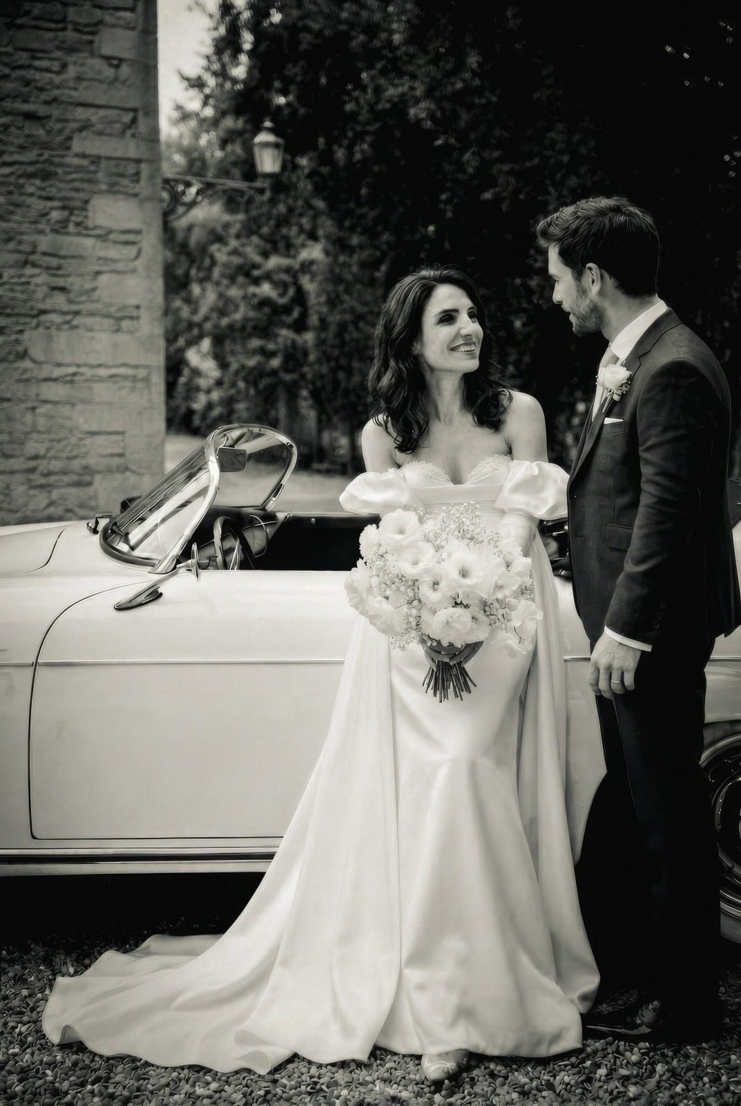 Bride in an elegant gown holds a bouquet while sharing a romantic moment with her groom beside a vintage car.