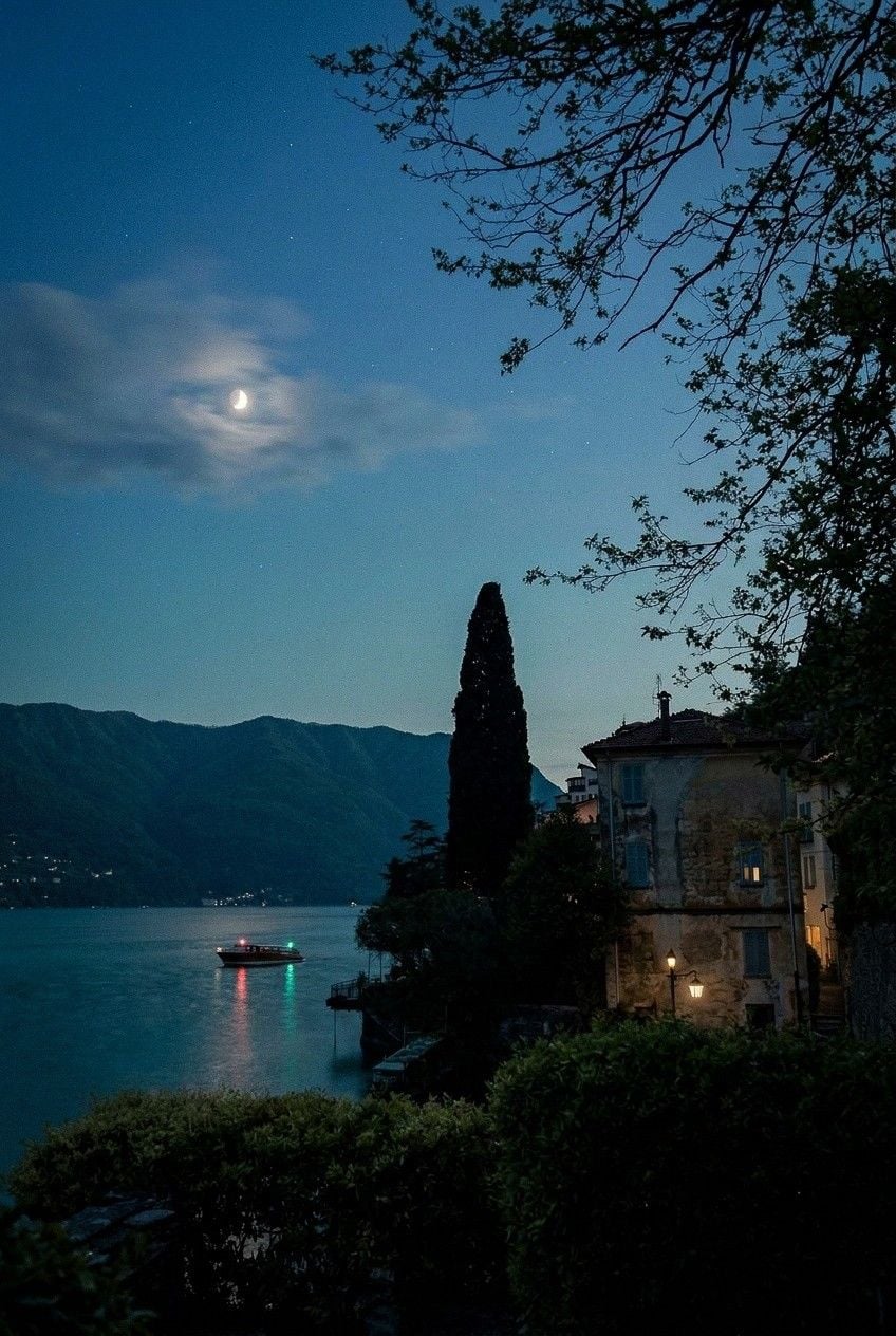 A serene lakeside scene at night, featuring a boat on calm waters, illuminated buildings, and a crescent moon above.