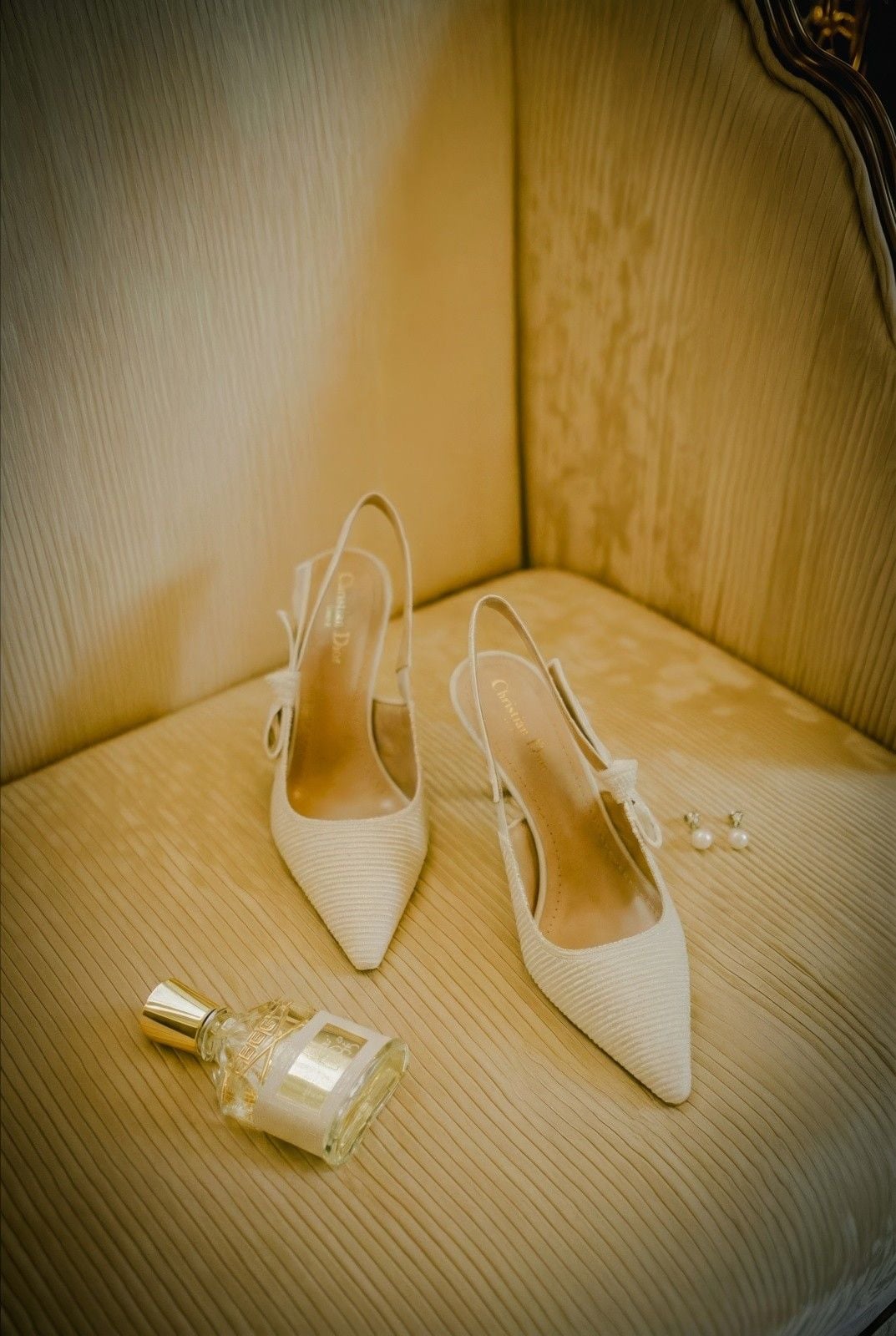 Elegant beige slingback heels paired with a bottle of perfume and pearl earrings on a plush, tufted chair.