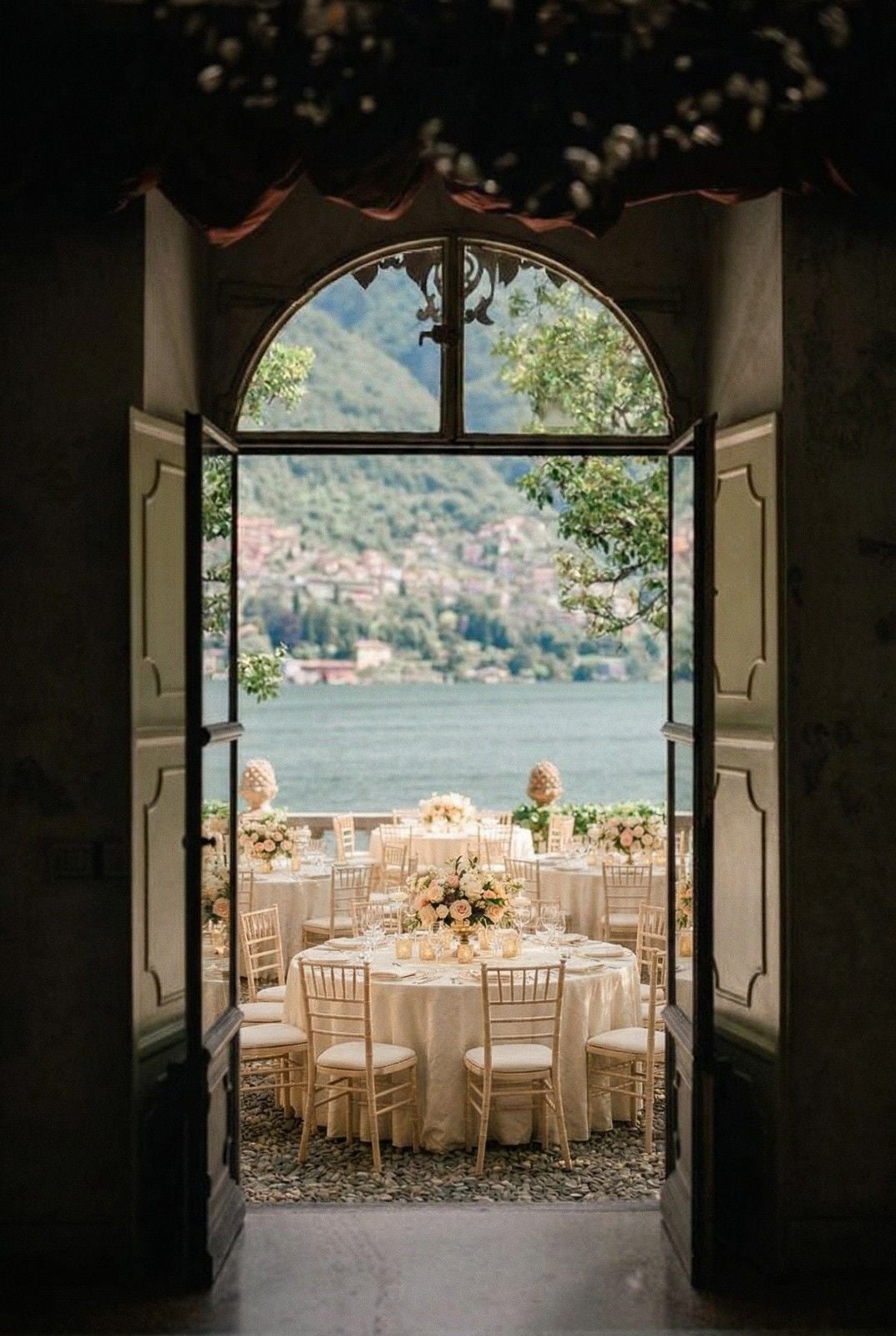 An elegant wedding setting with round tables adorned with floral centerpieces, framed by open doors and a lake view.