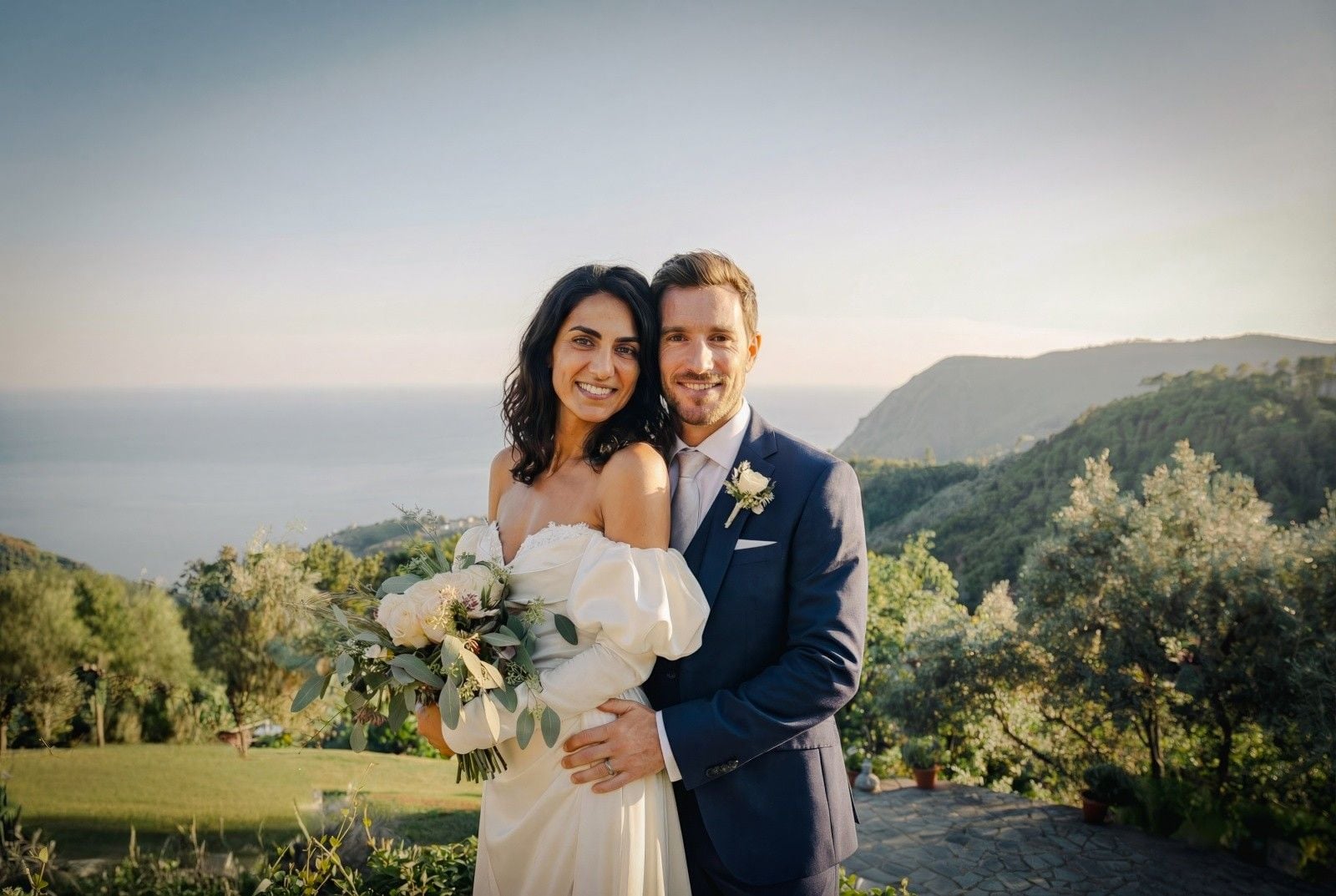 A joyful couple in formal wedding attire, surrounded by lush greenery and a stunning ocean backdrop.