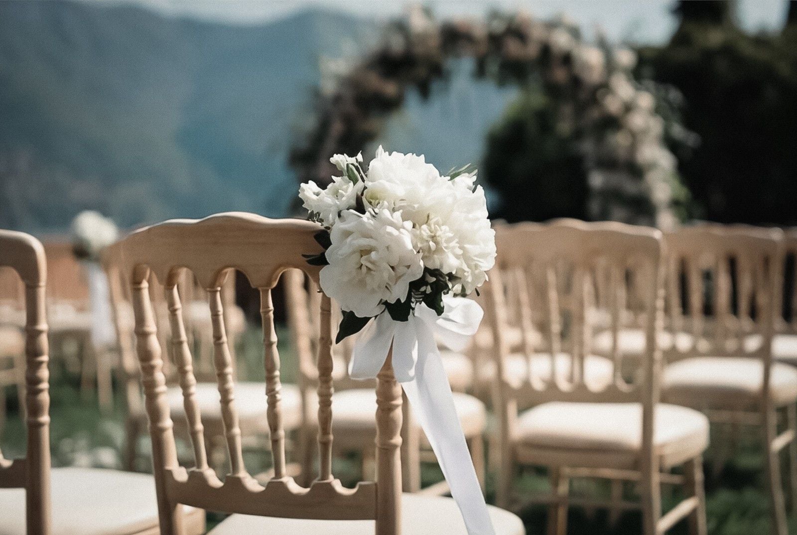 A serene outdoor wedding setup with wooden chairs and white floral decorations, set against a mountainous backdrop.