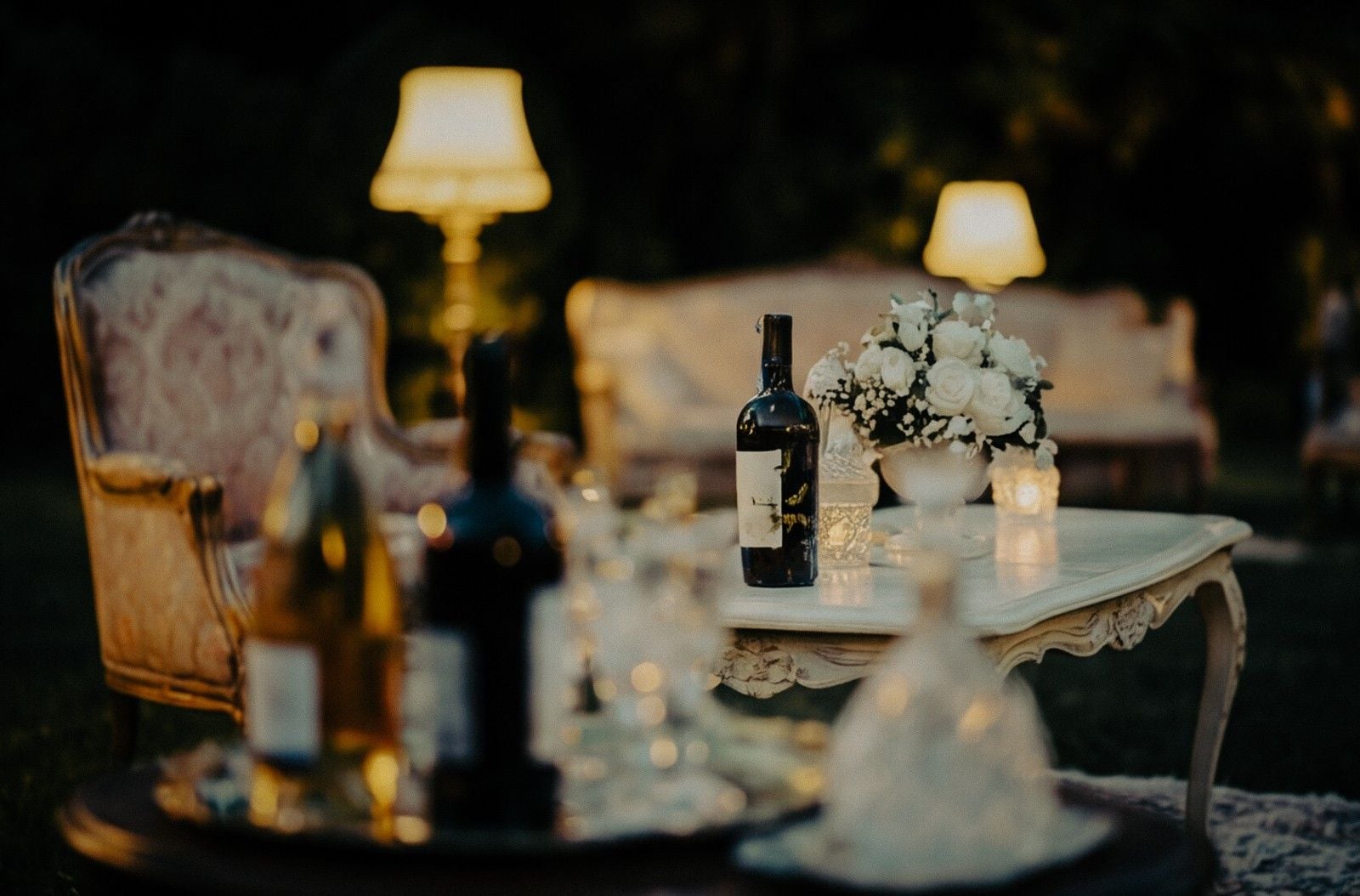 A romantic garden setting with elegant furniture, wine bottles, floral arrangements, and soft lighting creating a cozy ambiance.