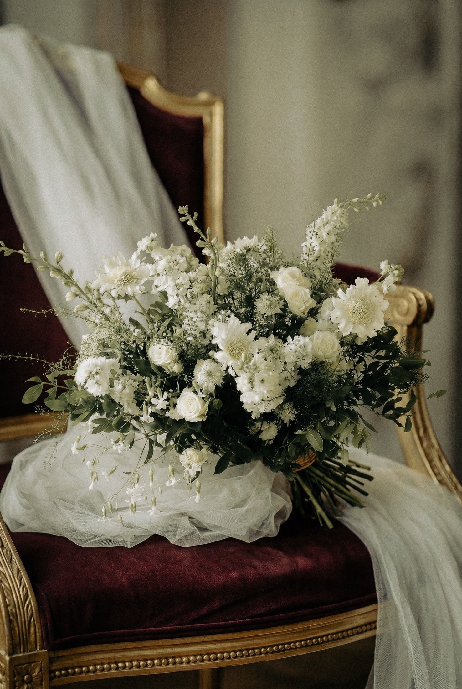 A beautiful bouquet of white flowers rests on a velvet chair, elegantly draped with sheer fabric, evoking romance.