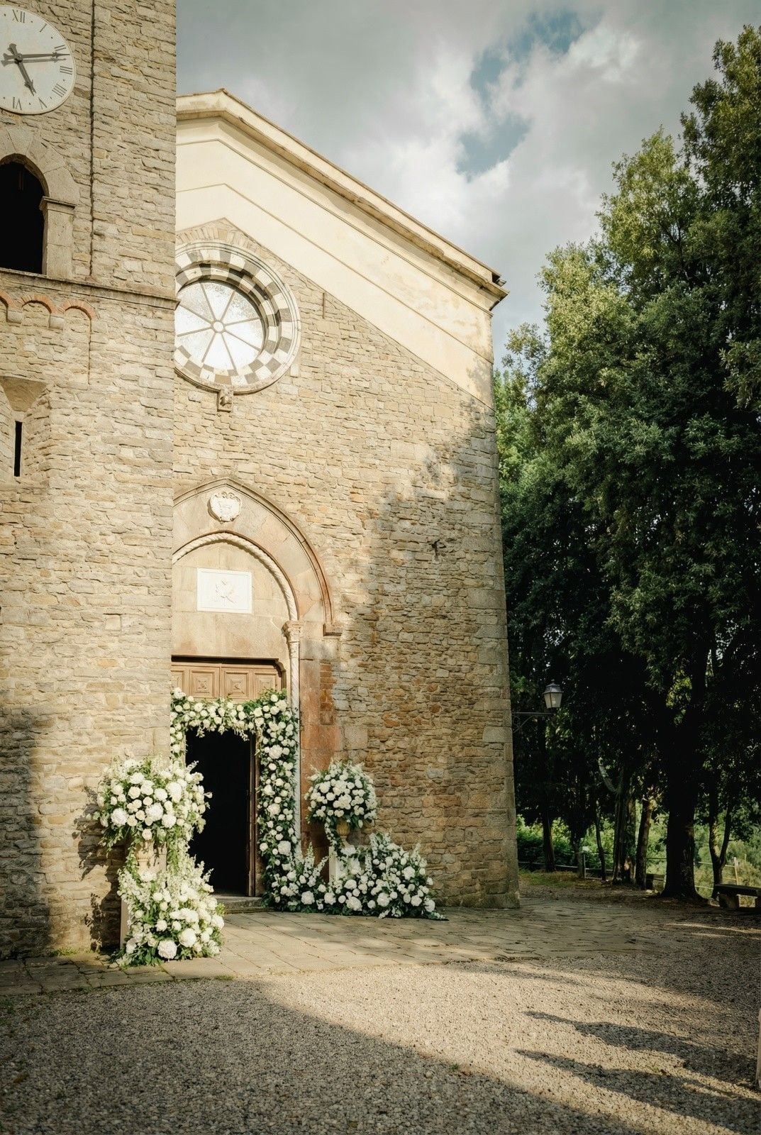 Stone church entrance adorned with lush white floral arrangements and greenery, set against a serene sky.