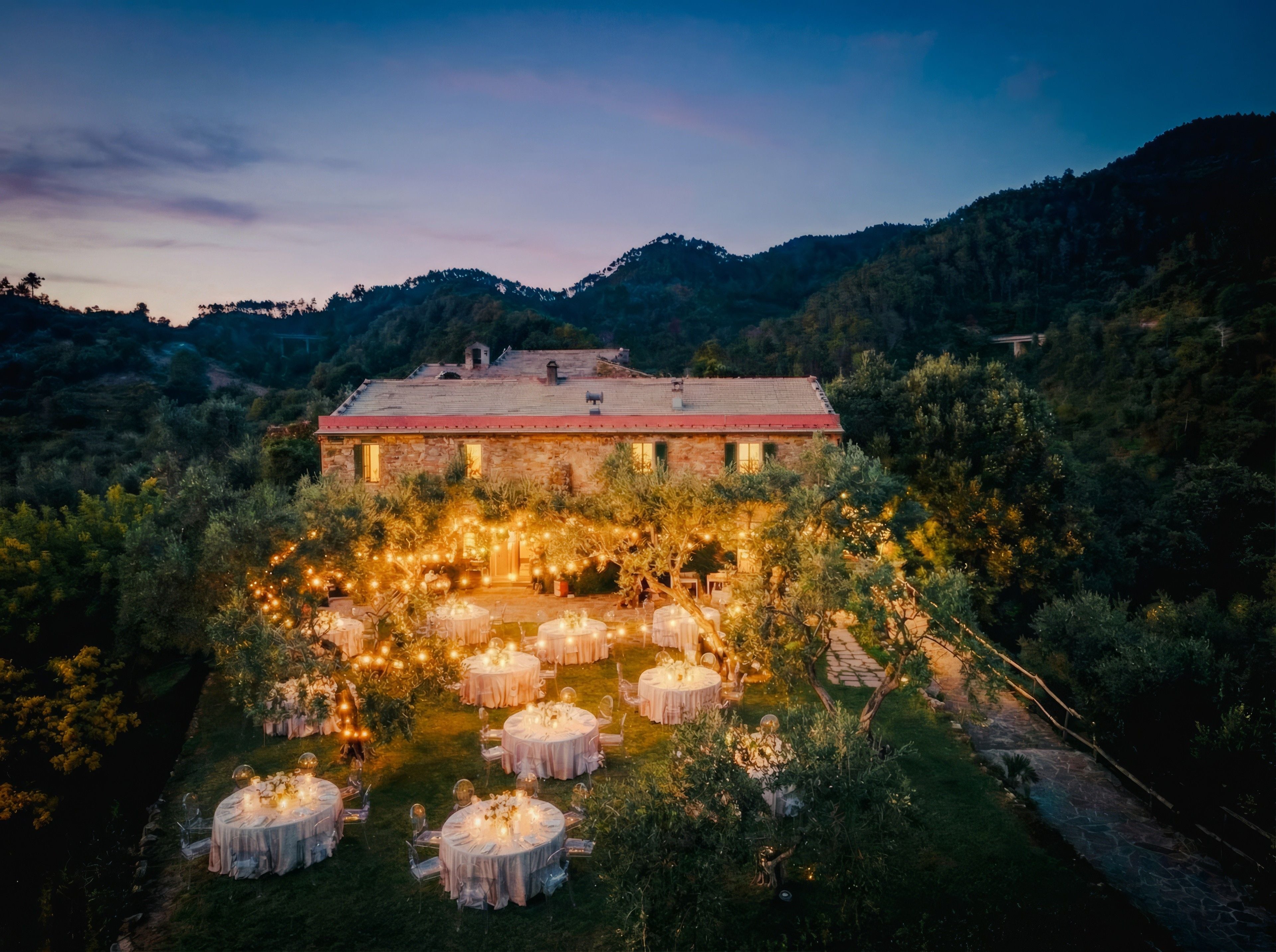 Elegant outdoor evening setup with round tables, twinkling lights, and a scenic hillside backdrop.