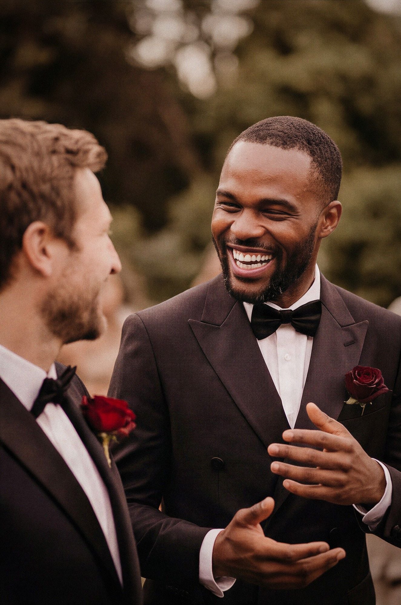 Two men in formal attire laugh joyfully during a wedding, with rose boutonnieres accenting their suits.