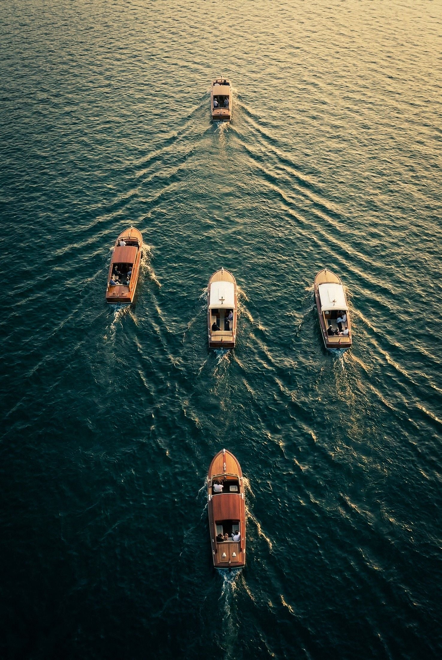 Six boats glide across calm water at sunset, creating gentle ripples under a warm golden light.