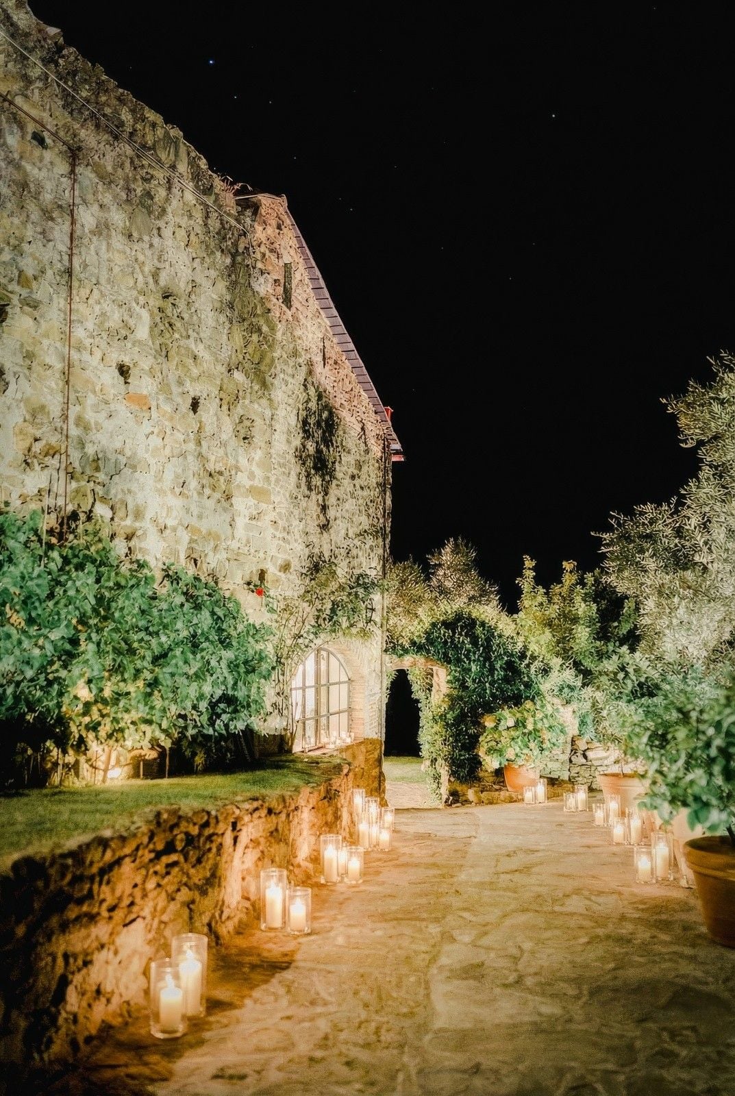 Warm candles illuminate a stone pathway leading to a rustic villa, surrounded by lush greenery under a night sky.