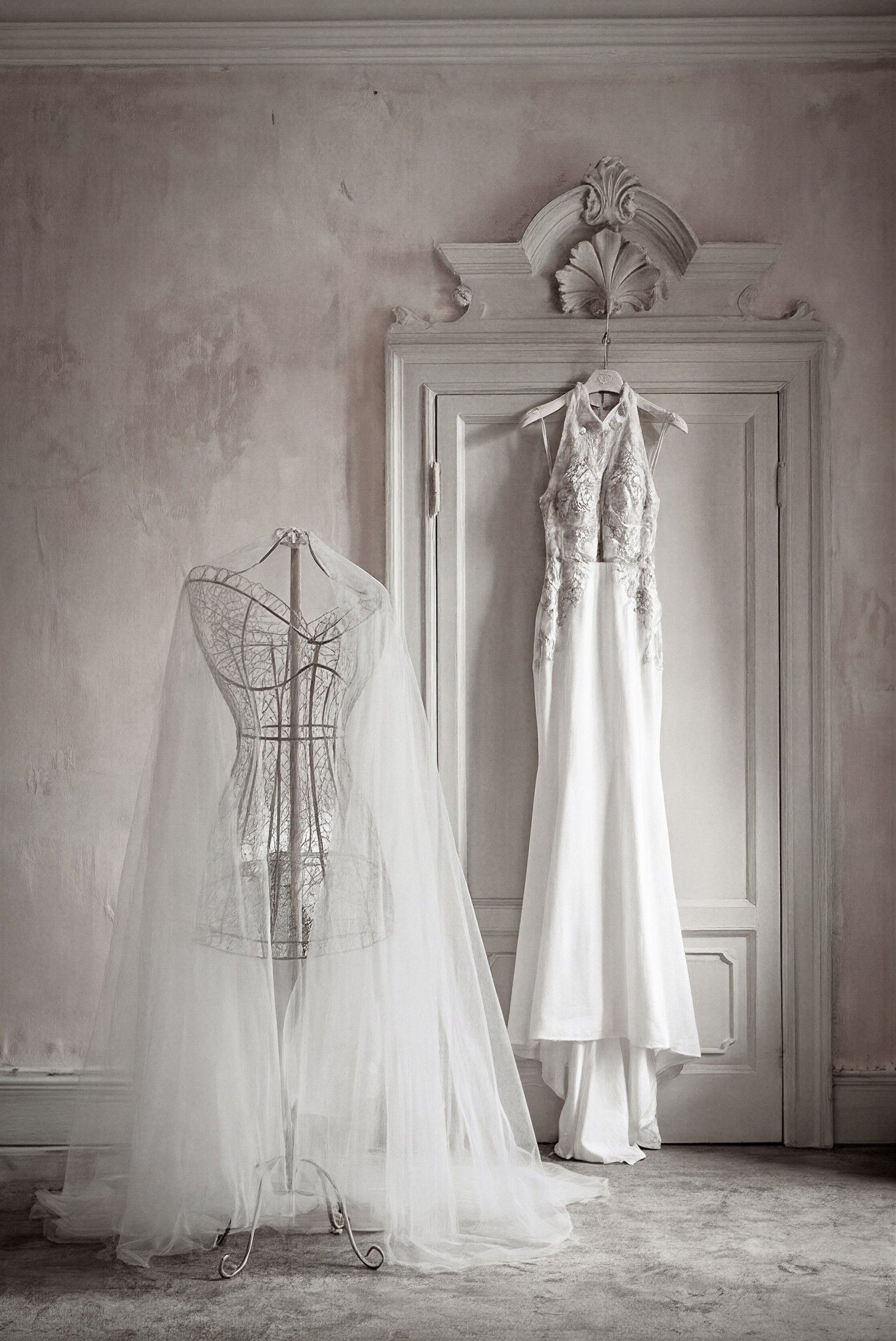 A delicate wedding gown hangs gracefully beside a lace-trimmed veil on a vintage dress form in a softly lit room.