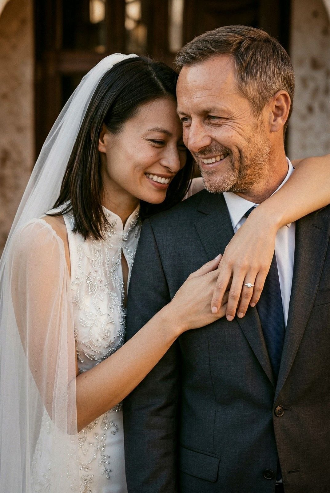 A joyful bride in a detailed white dress embraces a groom in a suit, both smiling in a warm, outdoor setting.