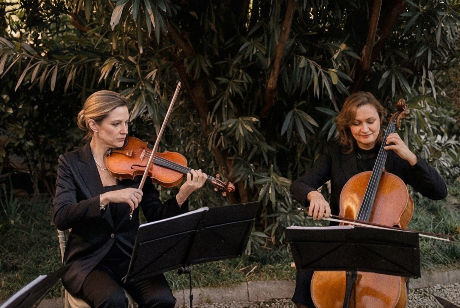 Two musicians play violin and cello outdoors amidst lush greenery, dressed elegantly for a wedding celebration.