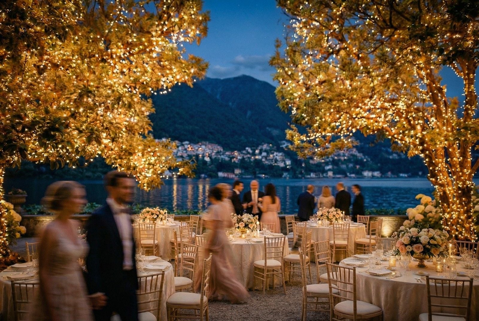 A beautifully lit outdoor wedding reception by a lake, featuring elegant tables, floral arrangements, and dancing guests.
