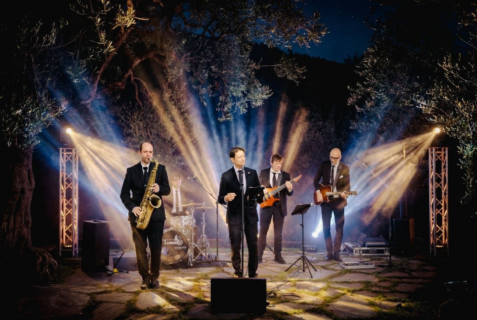 A live band performs in an outdoor setting, illuminated by dramatic lighting amidst olive trees.
