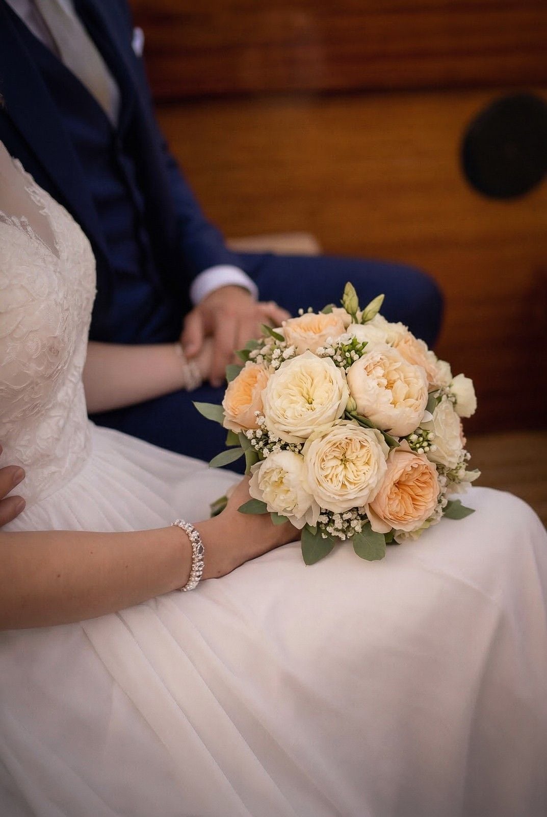 A bride holds a beautiful bouquet of soft peach and white roses while sharing a tender moment with her partner.