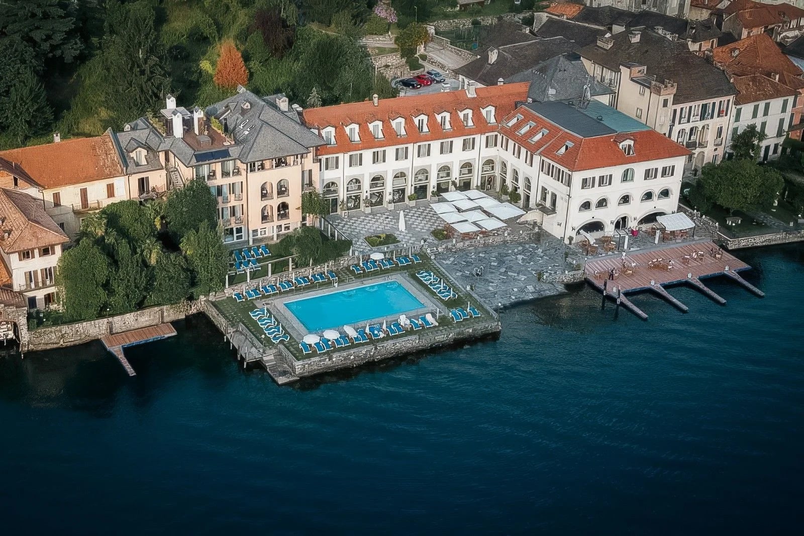 Luxury hotel by the lake featuring a pool, sun loungers, and elegant architecture surrounded by lush greenery.
