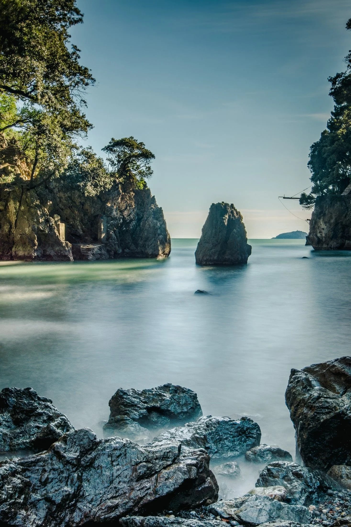 Serene coastal scene featuring smooth water, rocky formations, and lush greenery under a clear sky.