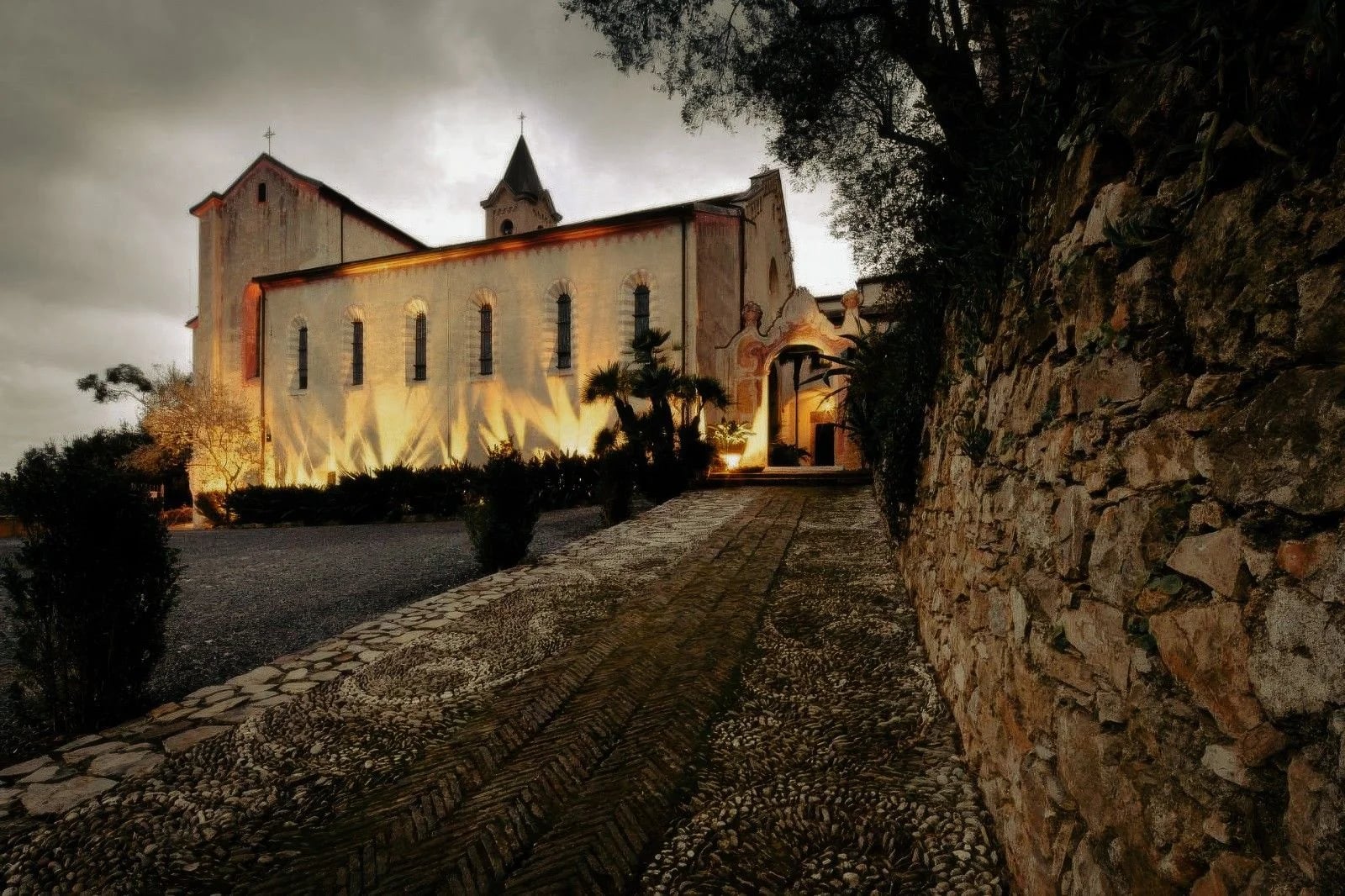 Historic church illuminated at dusk, surrounded by lush greenery and a textured stone pathway.