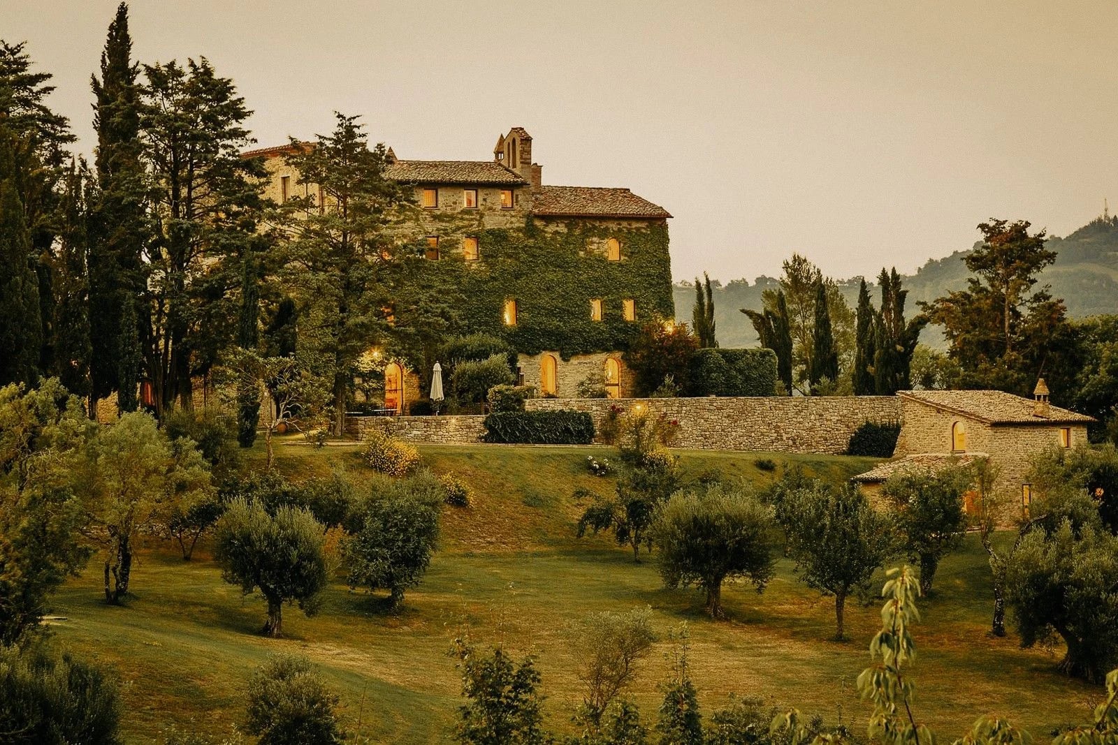 A rustic villa surrounded by lush greenery and trees, illuminated softly in the evening light.