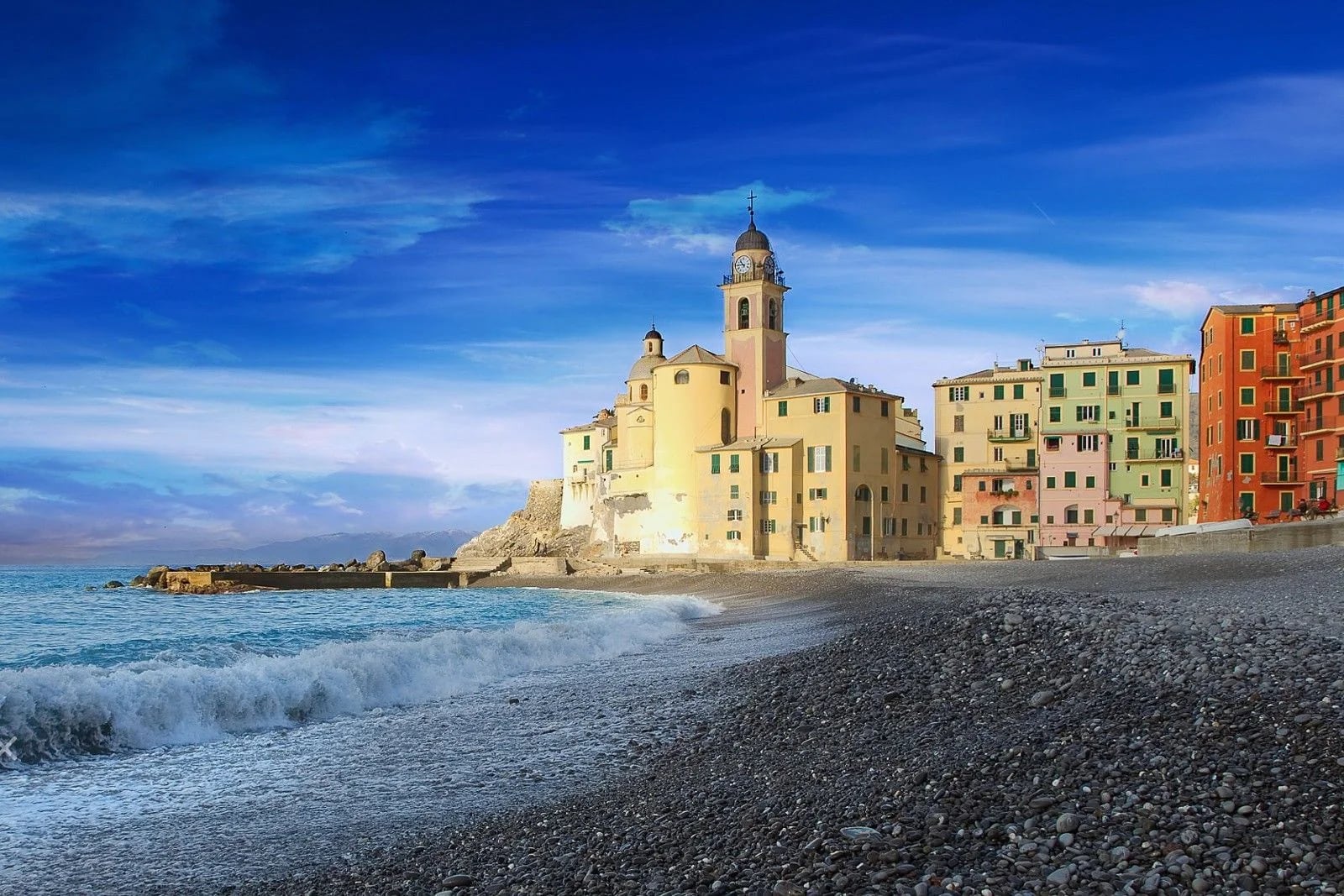 Colorful buildings line the coast of a Mediterranean village, with waves lapping at the pebbled beach under a blue sky.