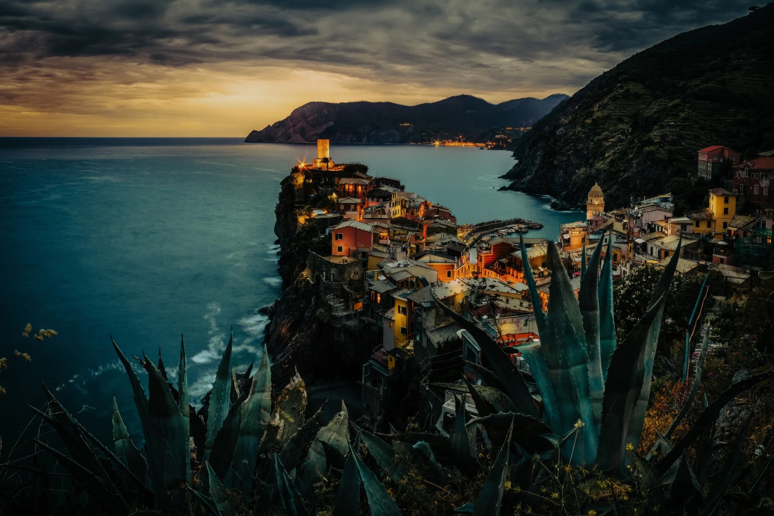 Colorful cliffside village of Vernazza twinkling at dusk, overlooking the sea with dramatic mountains in the background.