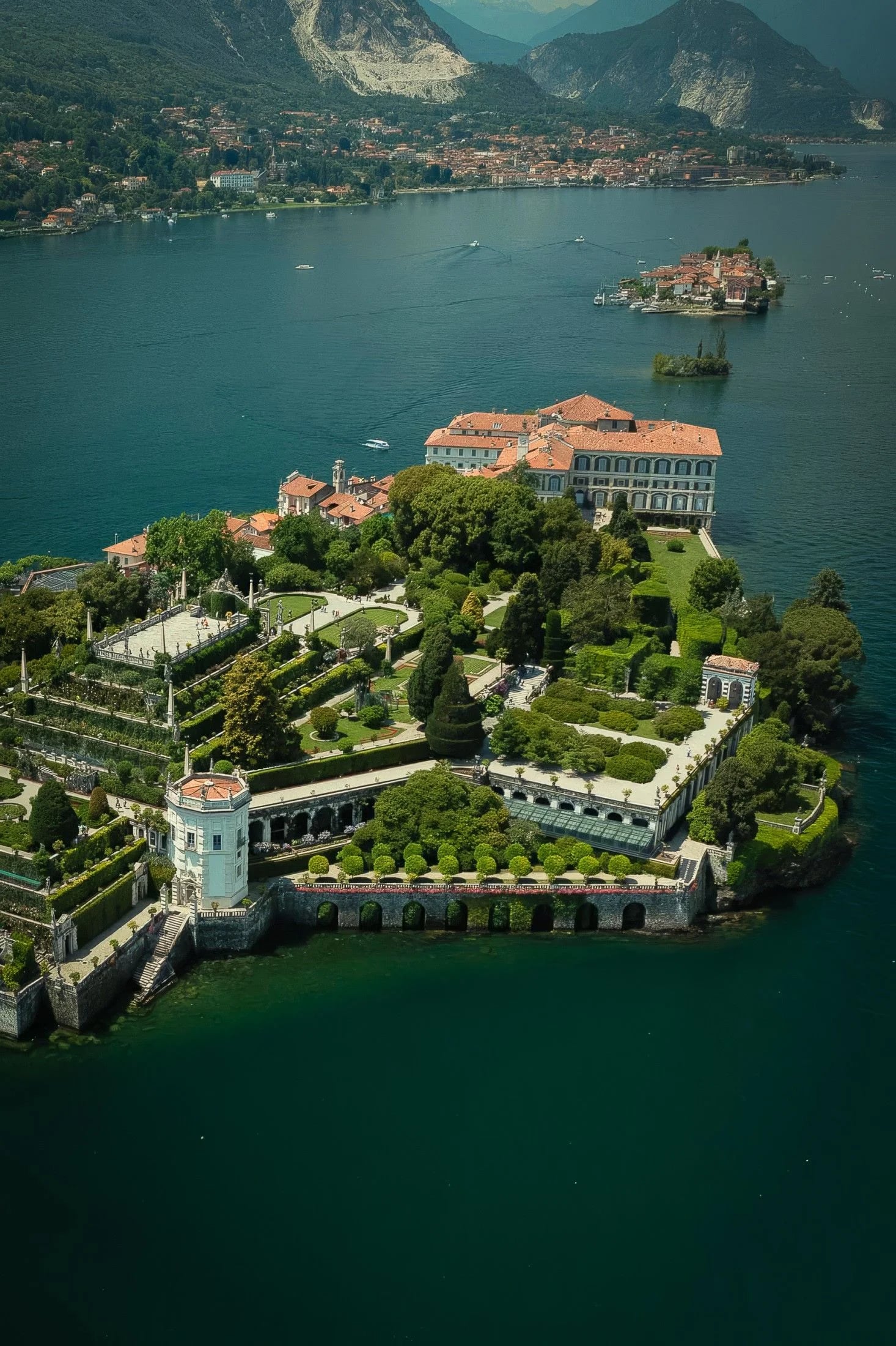 Aerial view of a grand villa surrounded by lush gardens and serene lake waters, with mountains in the distance.