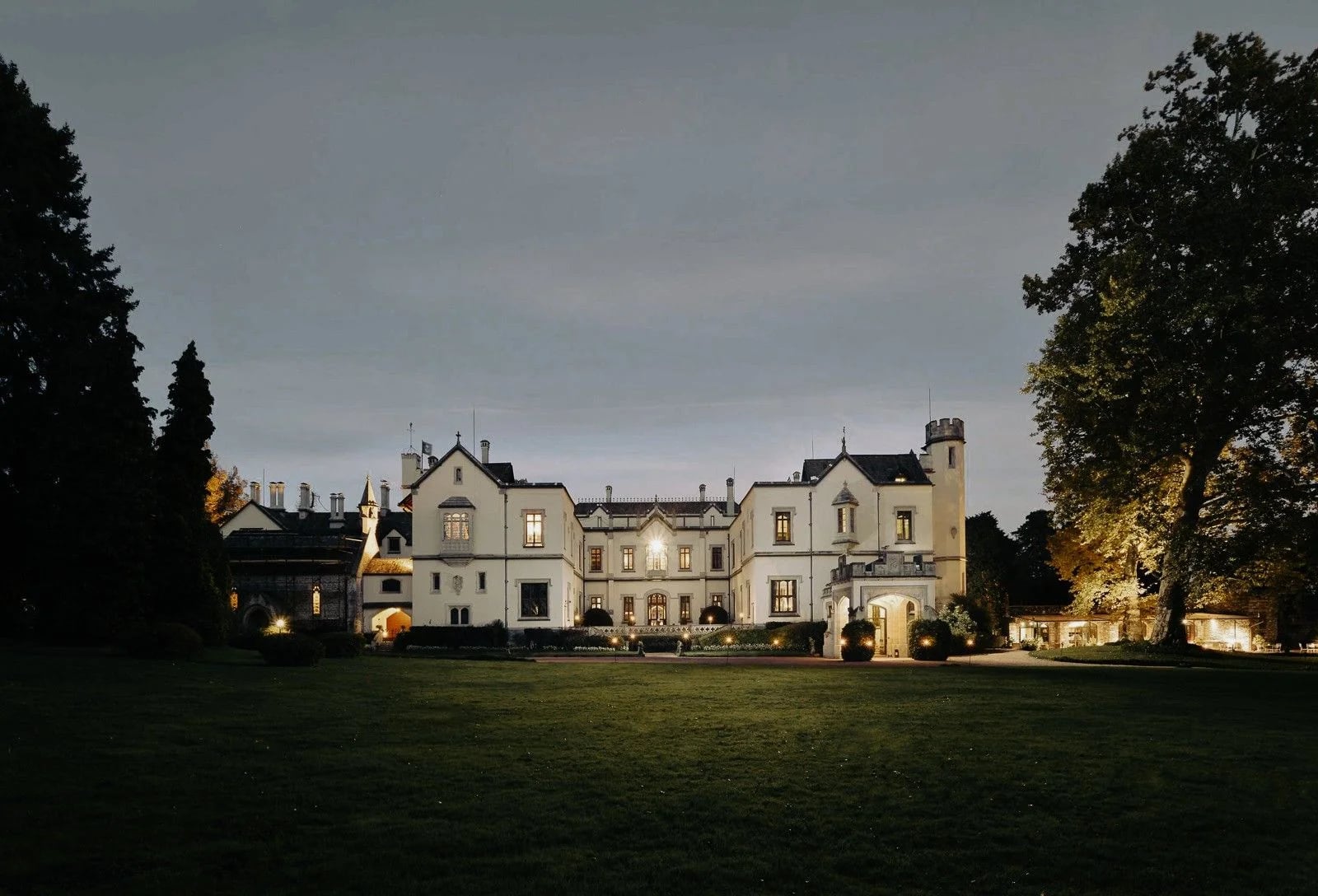Historic mansion illuminated at dusk, surrounded by lush greenery and elegant landscaping.