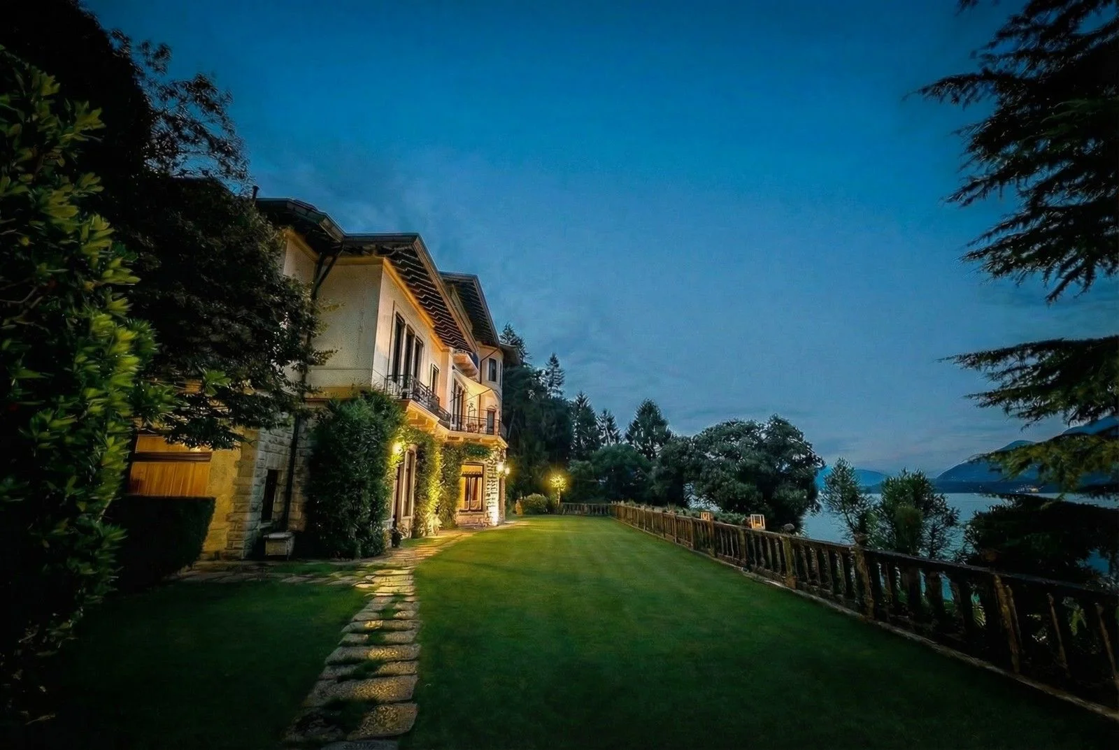 Elegant villa by a tranquil lake, illuminated softly at dusk, surrounded by lush greenery and a well-maintained garden.