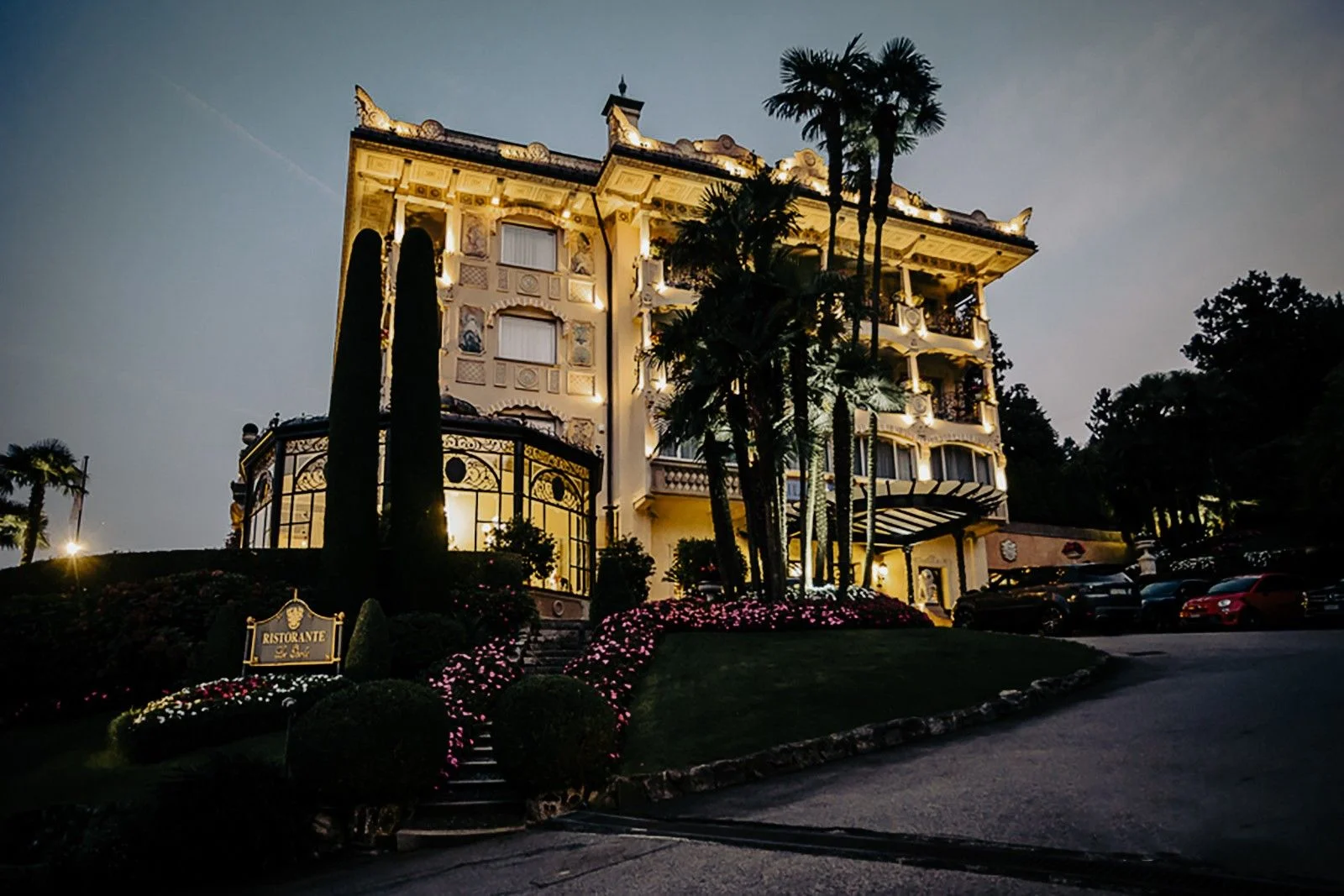 Luxurious mansion illuminated at dusk, surrounded by lush gardens and decorative plants.