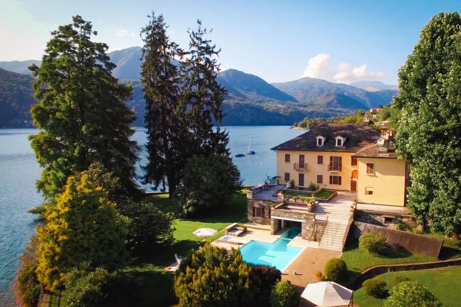 Scenic lakeside villa with a pool, surrounded by lush greenery and mountains under a clear blue sky.