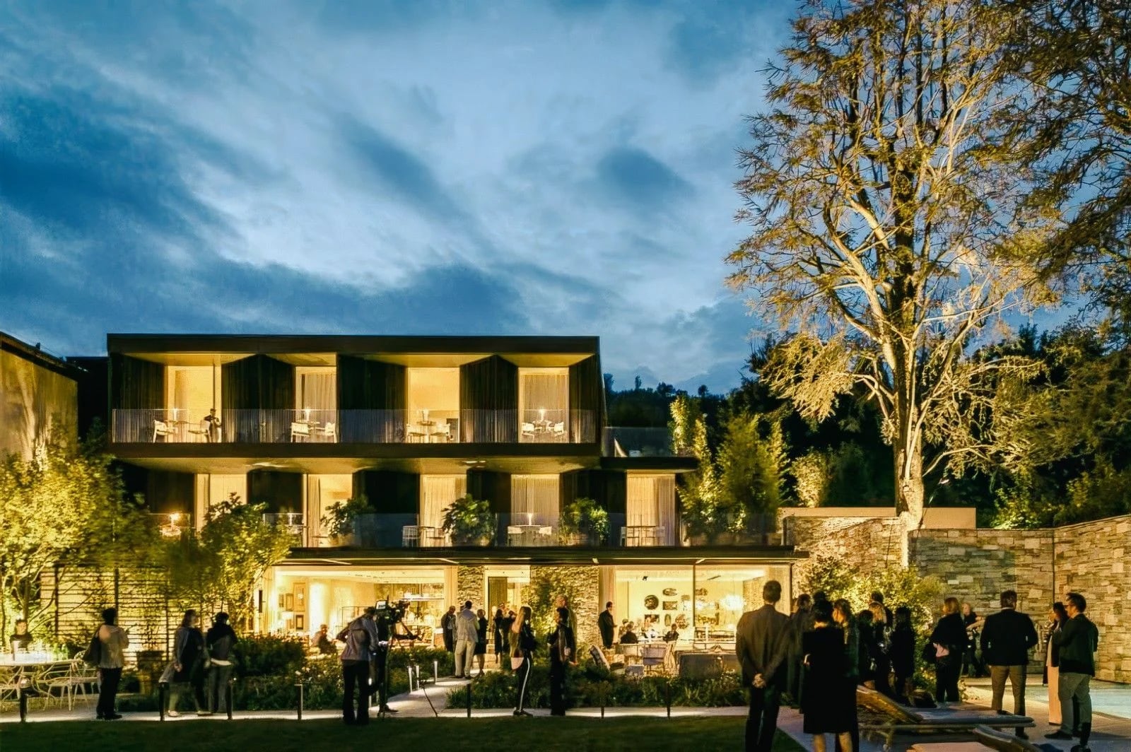 Elegant evening gathering outside a modern building surrounded by greenery, with guests socializing under a twilight sky.