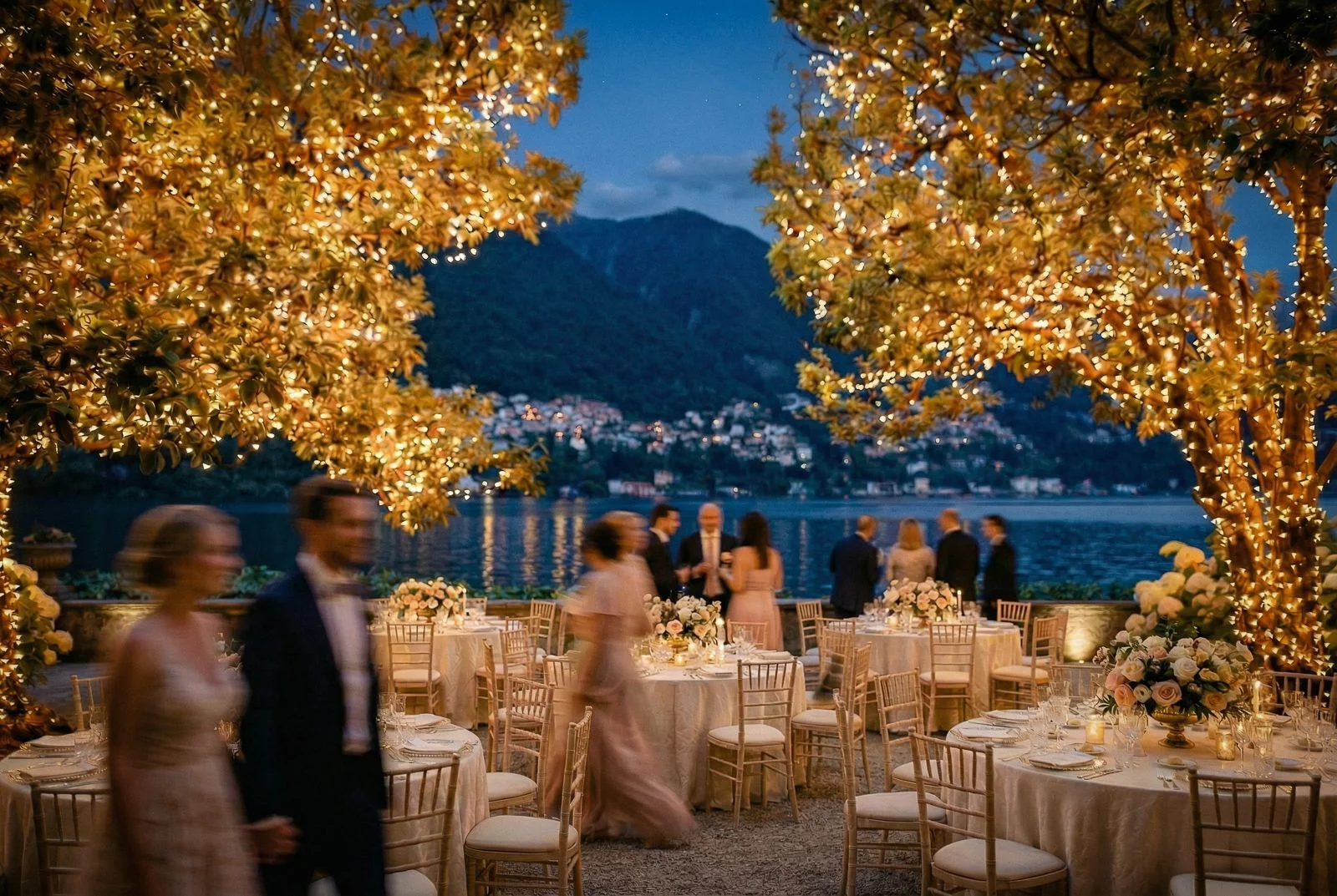 Outdoor wedding reception at dusk on Lake Como, fairy-lit trees framing candlelit tables and guests in motion