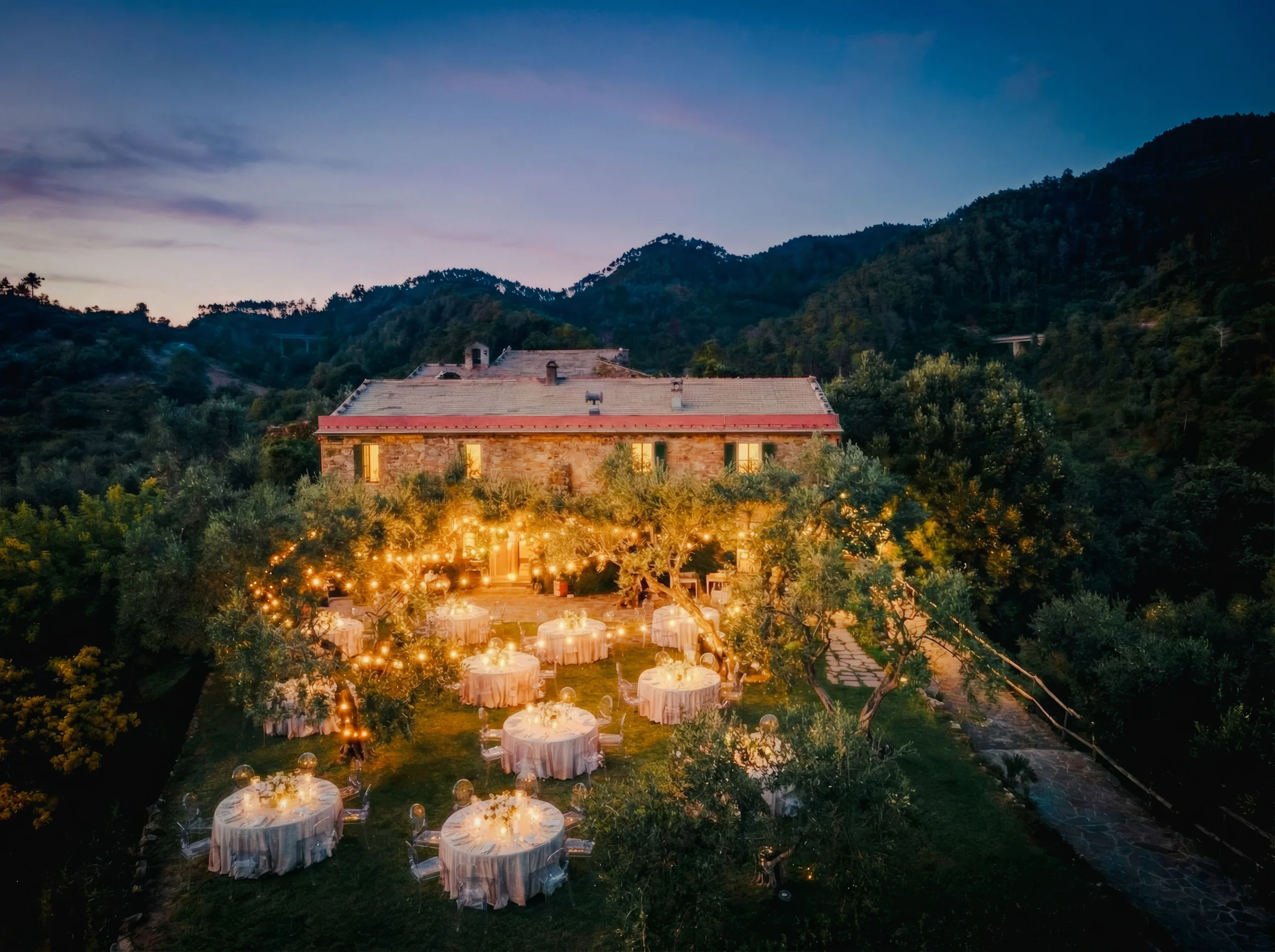 Elegant outdoor event set up with illuminated tables and string lights, surrounded by lush greenery and mountains at dusk.