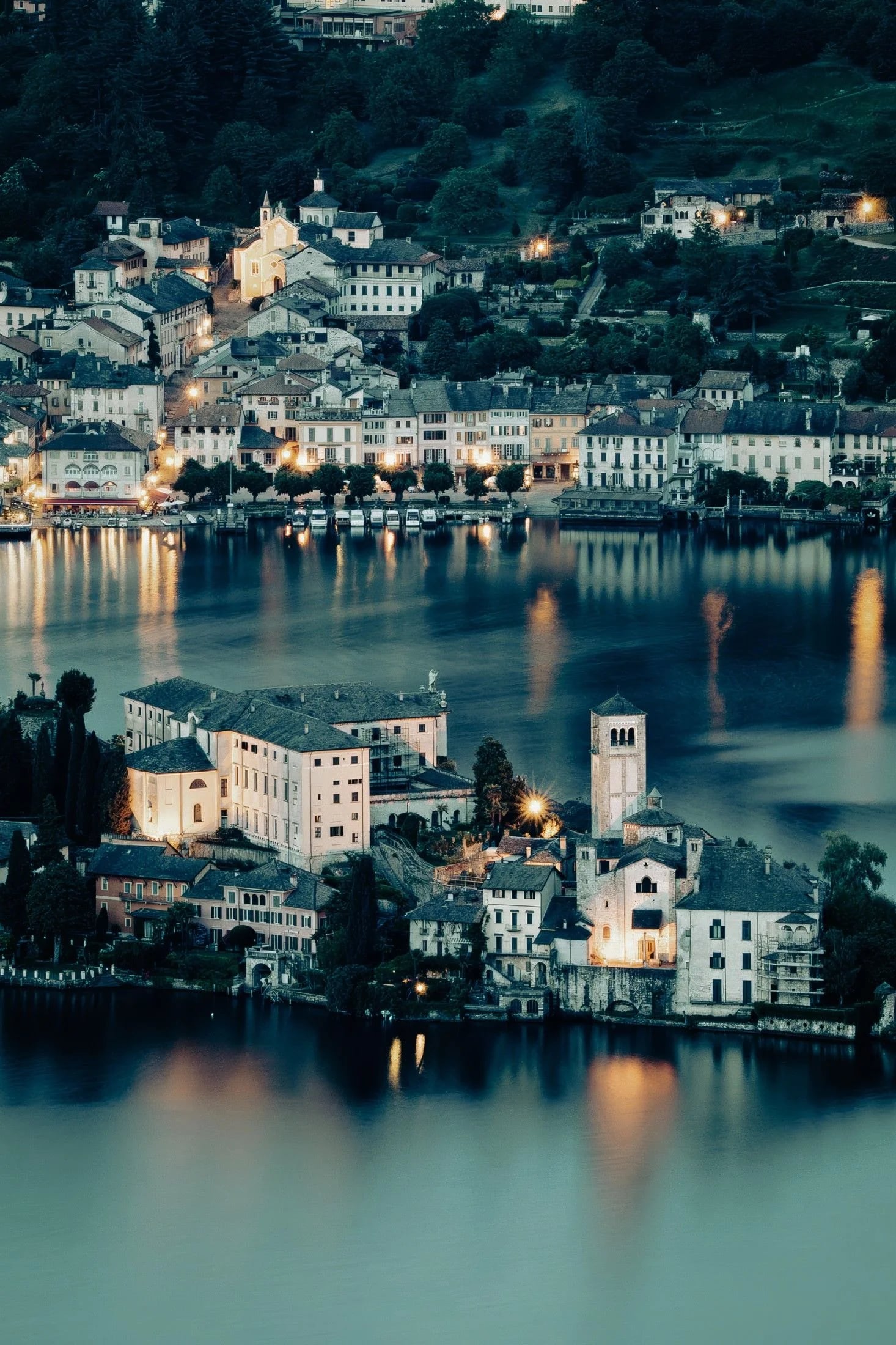 Picturesque lakeside village at twilight, reflecting lights on calm water, surrounded by serene hills and lush greenery.