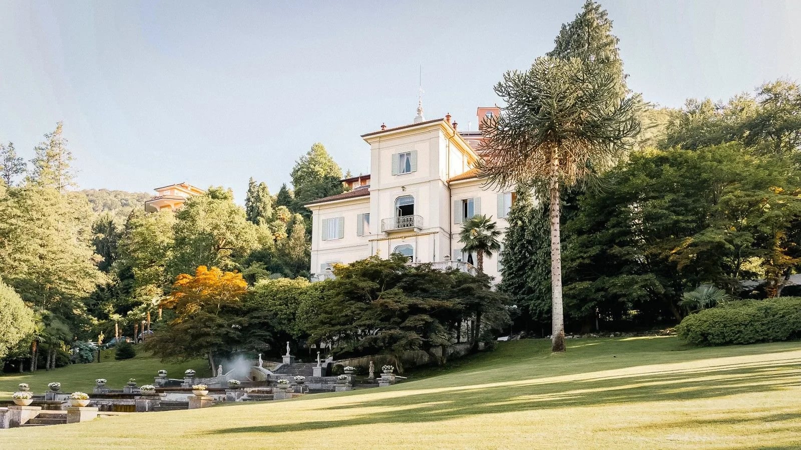 Elegant villa surrounded by lush greenery and tall trees, set against a clear blue sky.